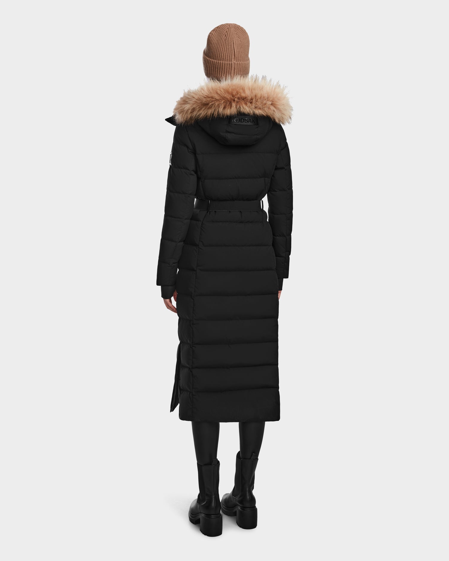 A person wearing the Rudsak ELSIE M - L123517 Black/Natural women’s maxi down puffer with a fur-trimmed hood, black boots, and a beige knit beanie stands facing away against a plain white background.