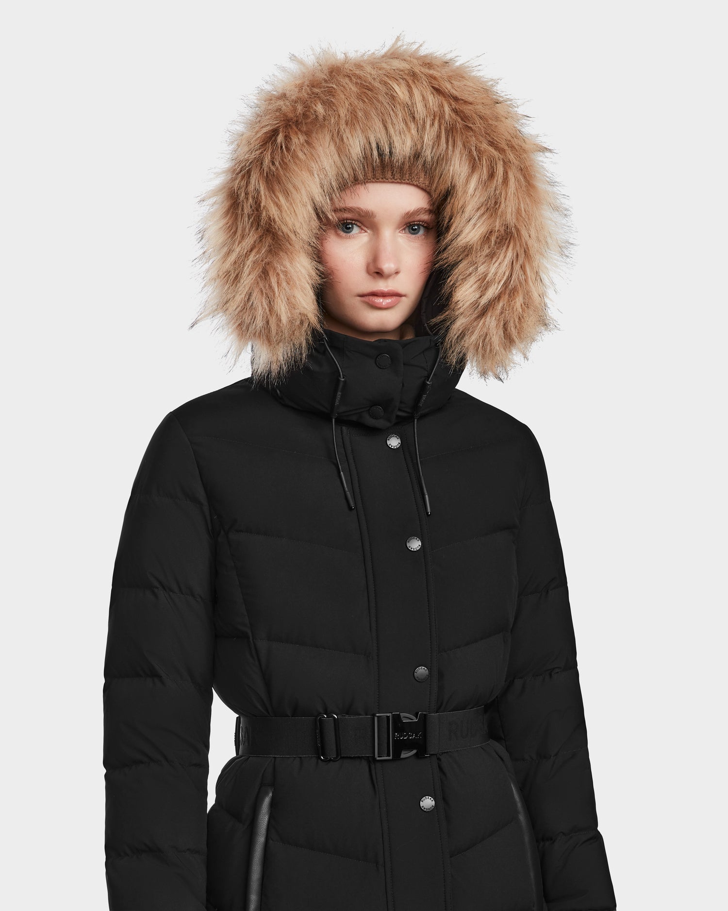 A person wears the Rudsak ELSIE M - L123517 Black/Natural women’s maxi down puffer coat with a large faux fur hood and RDS-certified down, standing against a plain light gray background.