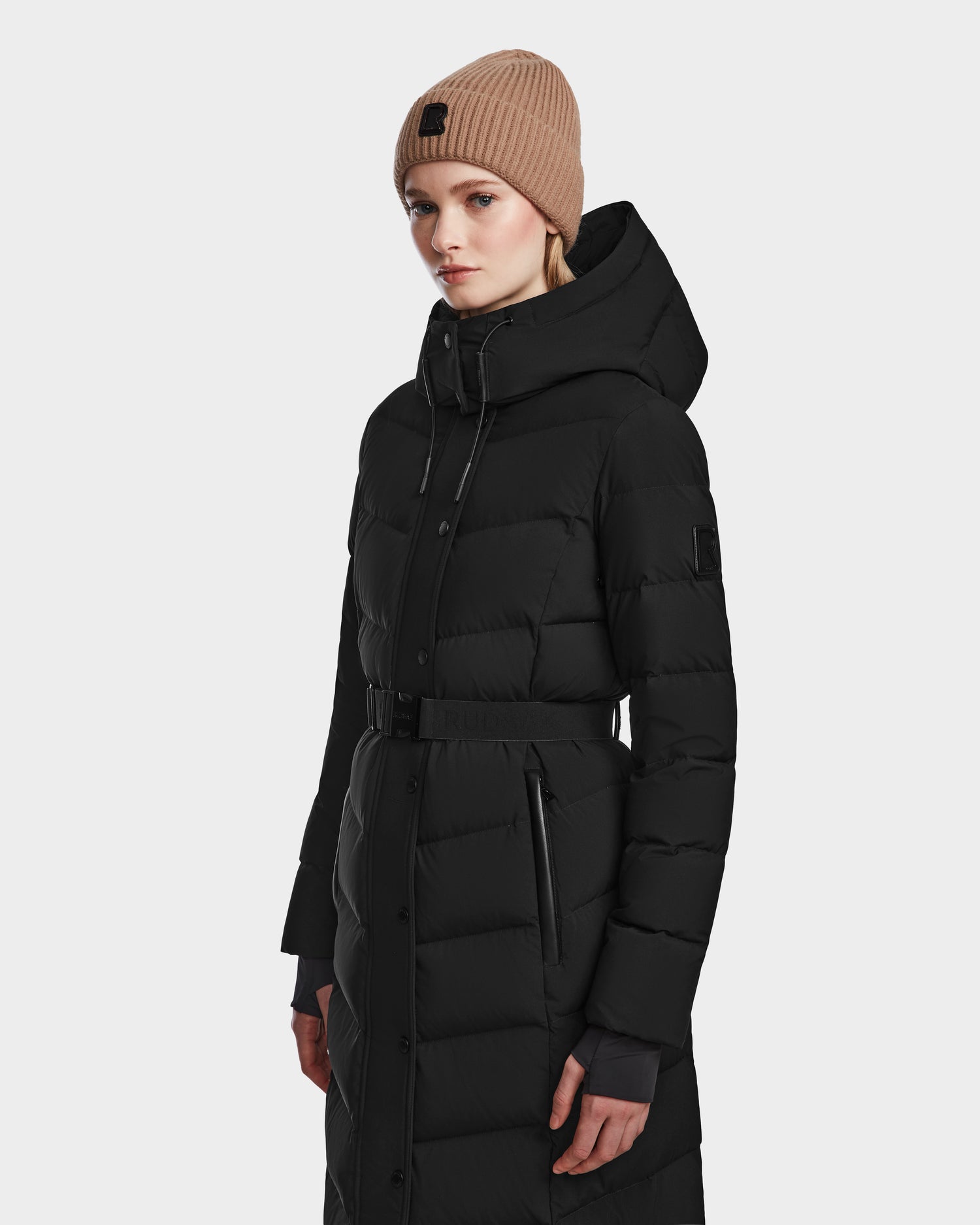 A woman in the Rudsak ELSIE M - L123517 Black/Natural maxi down puffer with a belted waist and RDS-certified down, paired with a beige knit beanie, stands against a light background, looking forward neutrally.