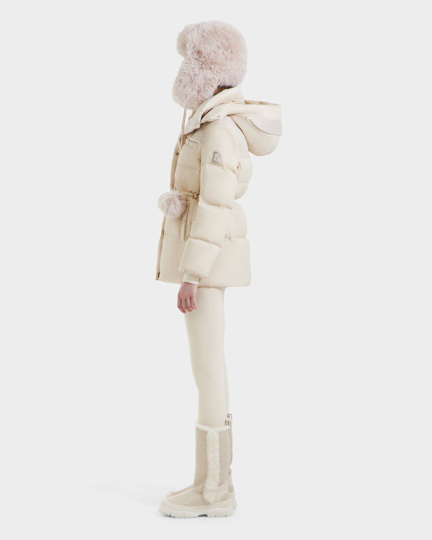 A person in profile wears the Rudsak EMBER M-M - L125524 Vanilla vegan-leather puffer jacket with a hood, paired with fitted pants, tall boots, and a large fluffy hat. Their all-cream winter outfit contrasts against a white background.