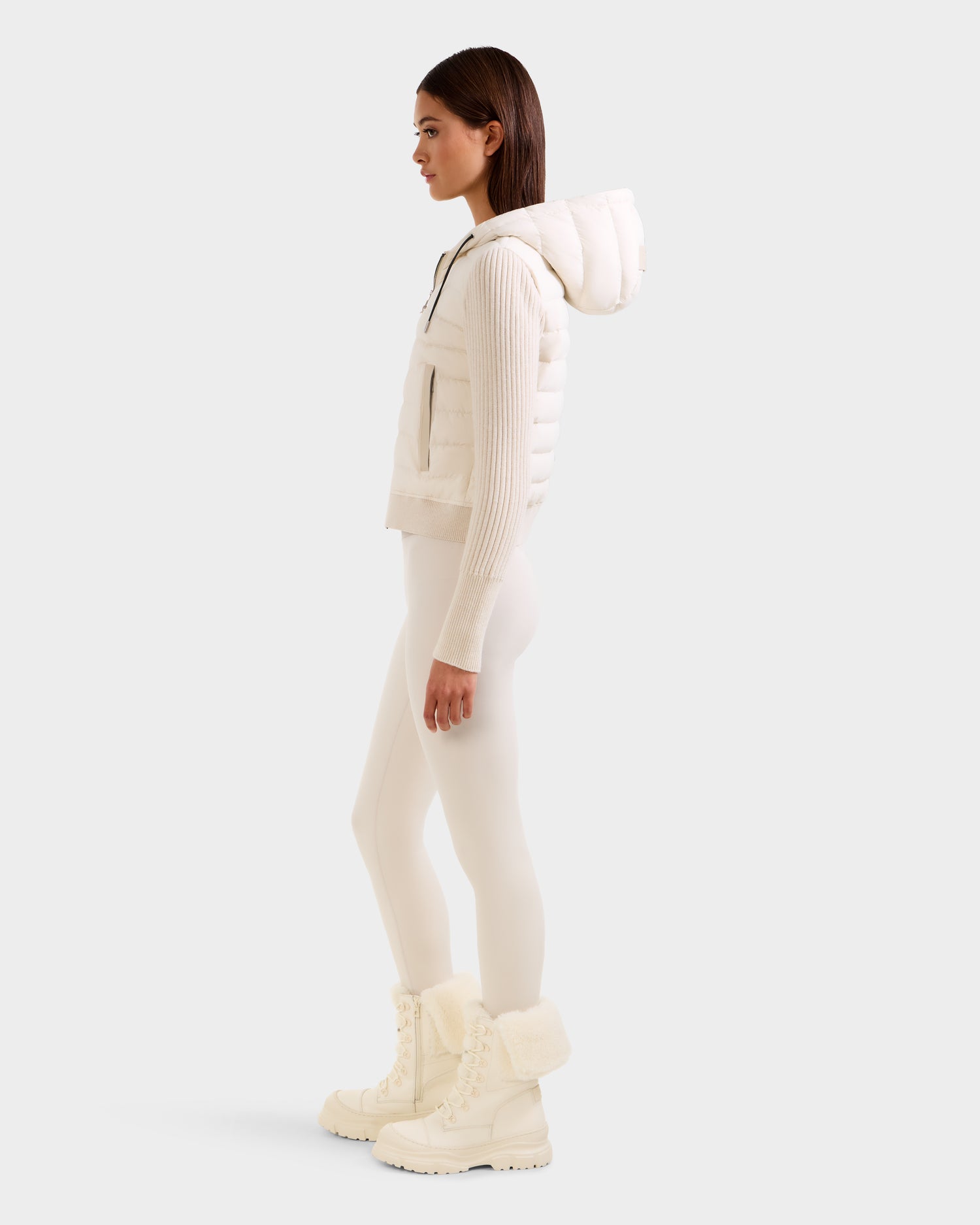A woman stands in profile against a light background wearing the Rudsak EMILY - L125515 Vanilla women’s hooded down bomber, paired with fitted leggings and fur-lined winter boots.