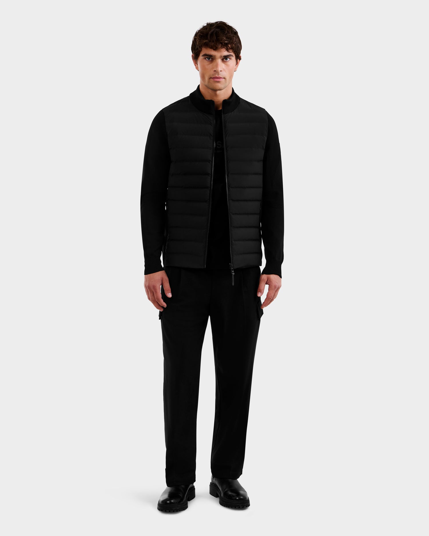 A man faces forward against a light gray background, wearing the Rudsak EMMETT - 6125029 Black mock-neck hybrid jacket with lightweight down insulation over a black shirt, black pants, and black shoes.