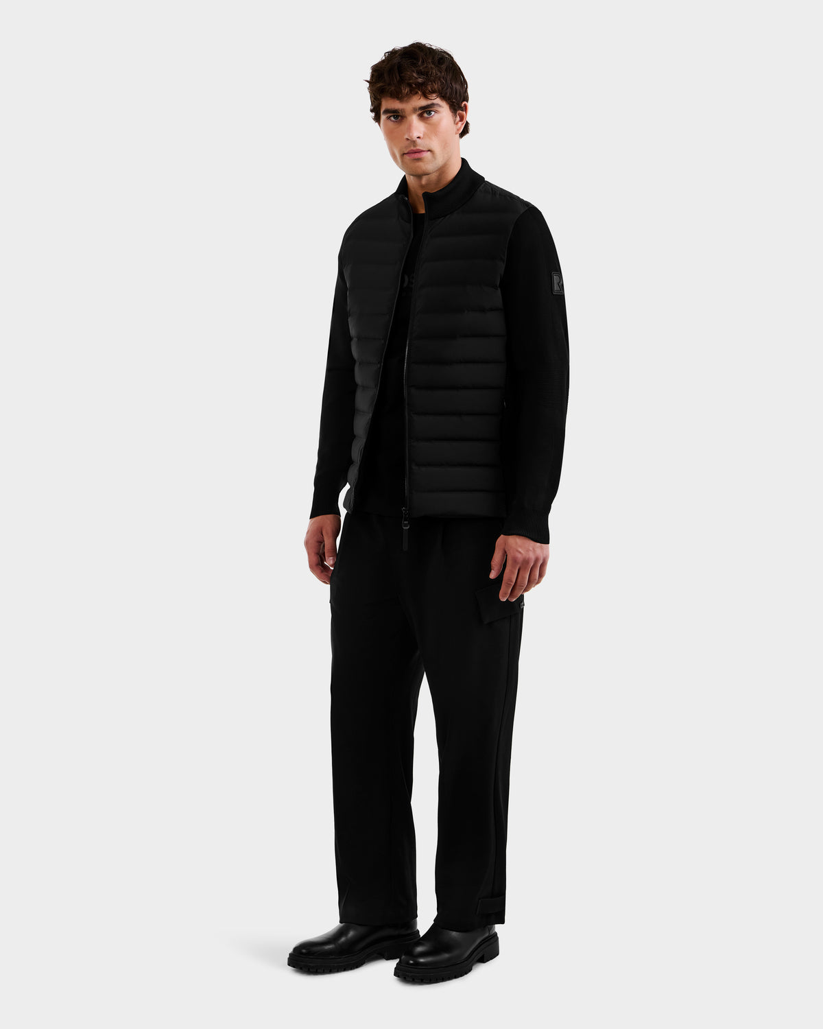 A man stands facing forward wearing the Rudsak EMMETT - 6125029 Black quilted vest, featuring lightweight down insulation, paired with a black long-sleeve shirt, black pants, and black shoes against a plain light background—a refined look for the modern men’s wardrobe.