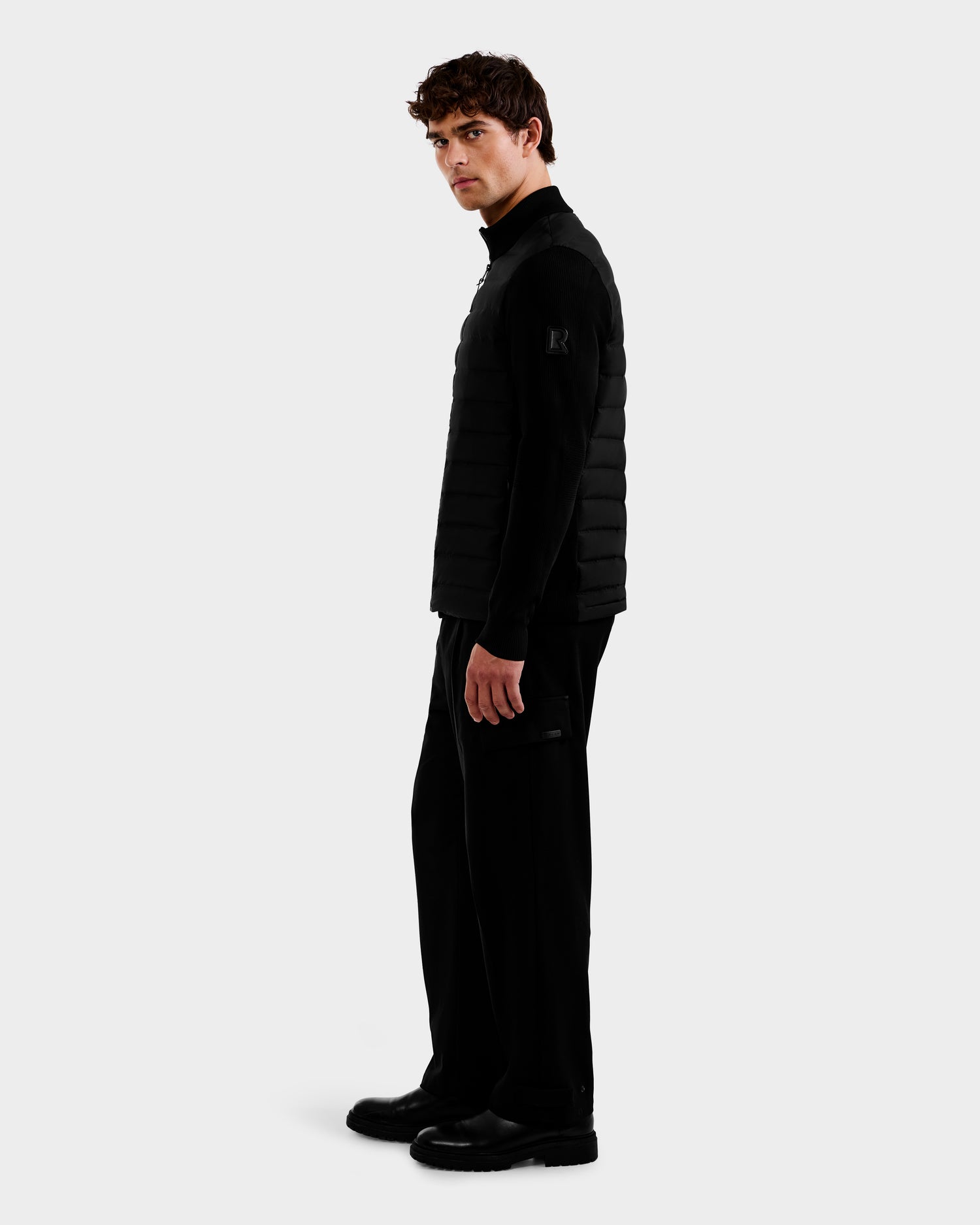 A man with short curly hair stands sideways, wearing the Rudsak EMMETT - 6125029 Black mock-neck hybrid jacket with lightweight down insulation, paired with black pants and shoes against a plain light background—modern menswear at its best.