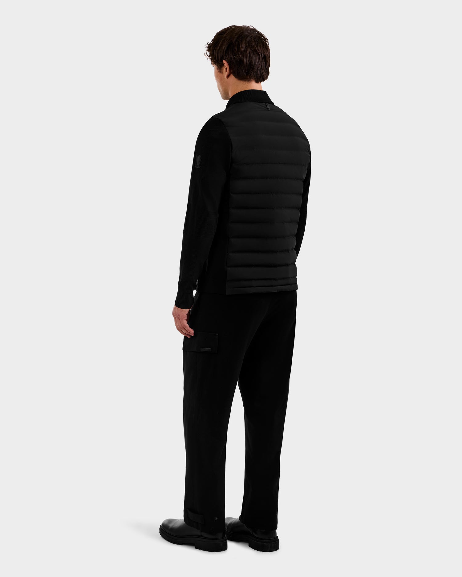 A person stands facing away, wearing the Rudsak EMMETT - 6125029 Black mock-neck hybrid jacket with lightweight down insulation, paired with black cargo pants and shoes, set against a plain light gray background.