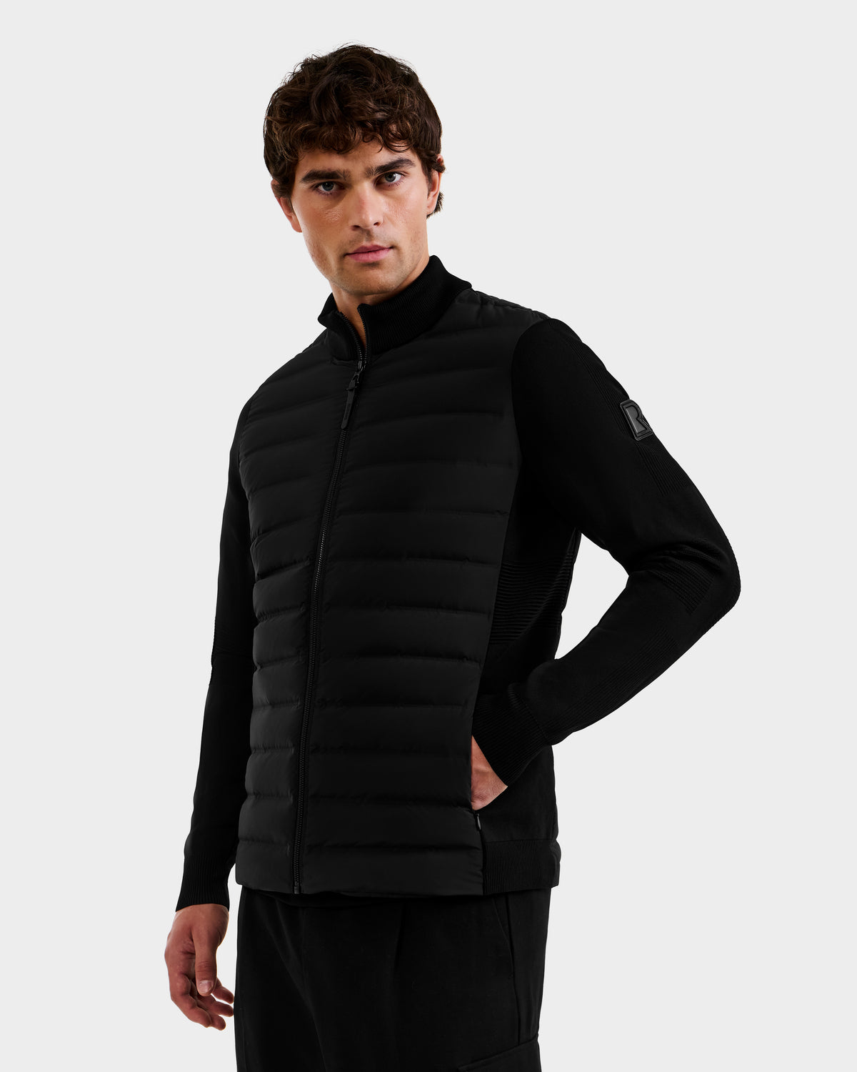 A man with short brown hair wears the Rudsak EMMETT - 6125029 Black mock-neck hybrid jacket, featuring quilted front panels, side pockets, and lightweight down insulation—an essential addition to the modern men’s wardrobe—as he stands against a plain white background.