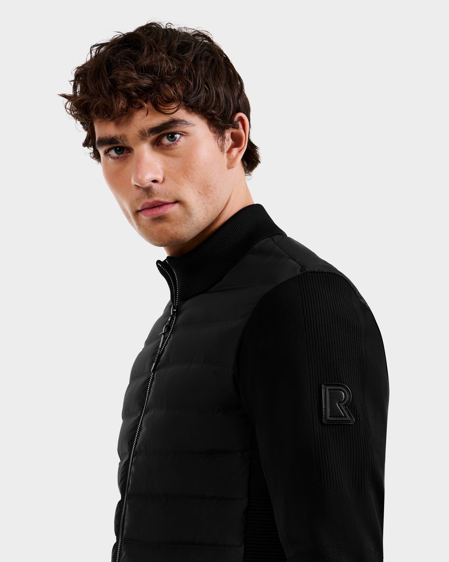 A young man with short curly hair models the Rudsak EMMETT - 6125029 Black jacket, featuring ribbed sleeves, lightweight down insulation, and an embossed R patch, as he glances over his shoulder against a plain light background.