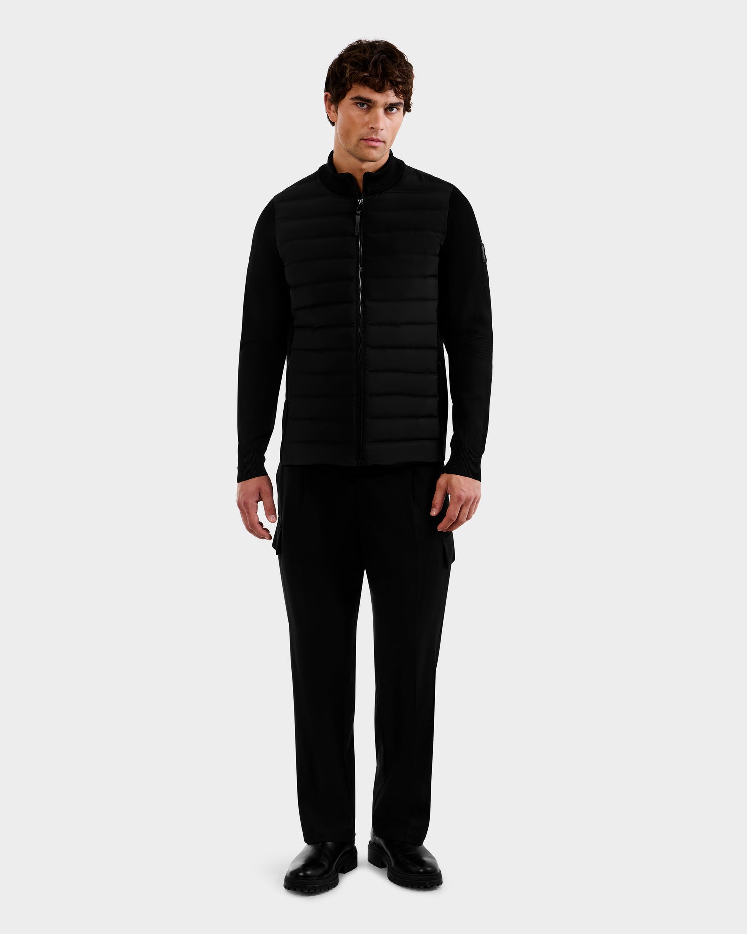 A man faces forward wearing the Rudsak EMMETT - 6125029 Black mock-neck hybrid jacket with lightweight down insulation, paired with black pants and shoes, against a plain light gray background.