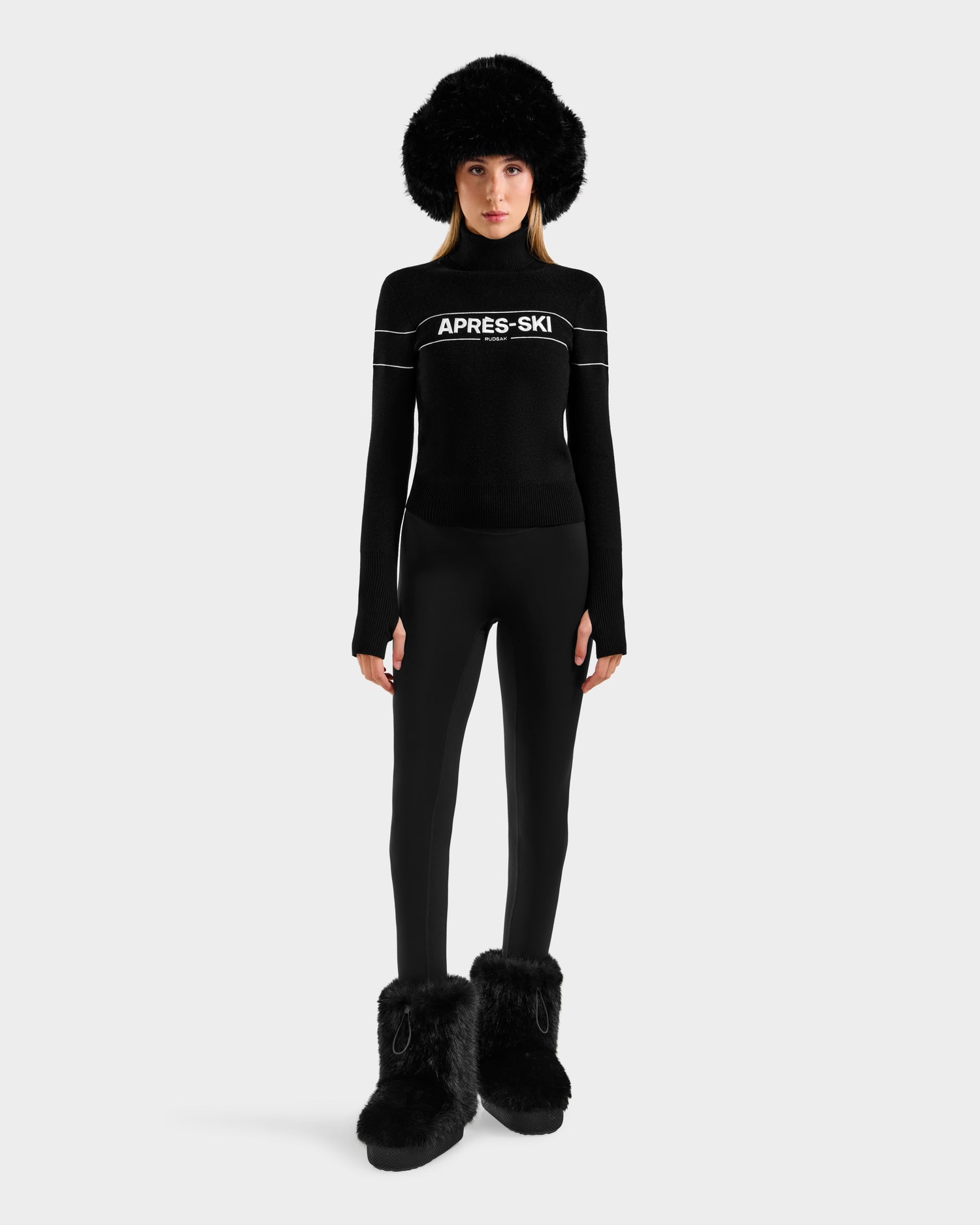 A person wearing Rudsak's ERIKA - 8523536 Black turtleneck sweater, black leggings, black furry boots, and a large black furry hat stands against a white background—perfect winter wardrobe inspiration.