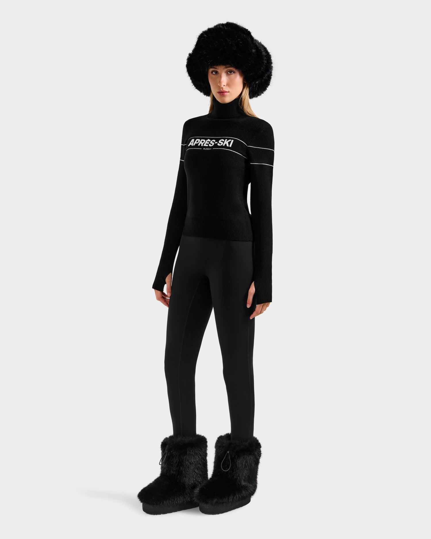 A woman wears the Rudsak ERIKA - 8523536 Black turtleneck sweater, paired with black fitted pants, fur boots, and a large fur hat against a white backdrop—a must-have winter look.