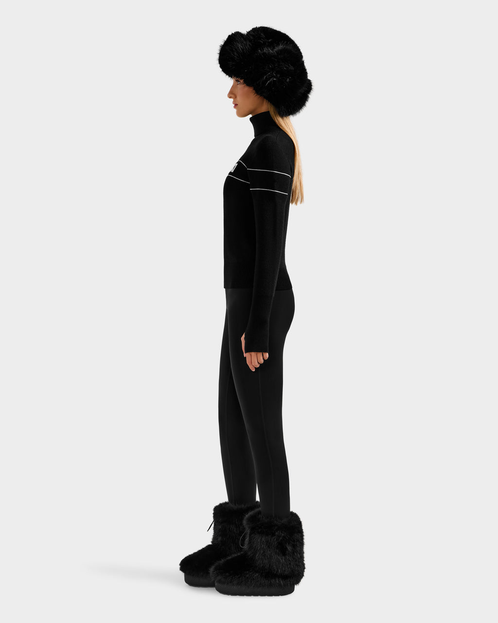 A woman stands in profile wearing the Rudsak ERIKA - 8523536 Black fur hat, an après-ski turtleneck with white stripes, black leggings, and black fur boots against a plain light background.