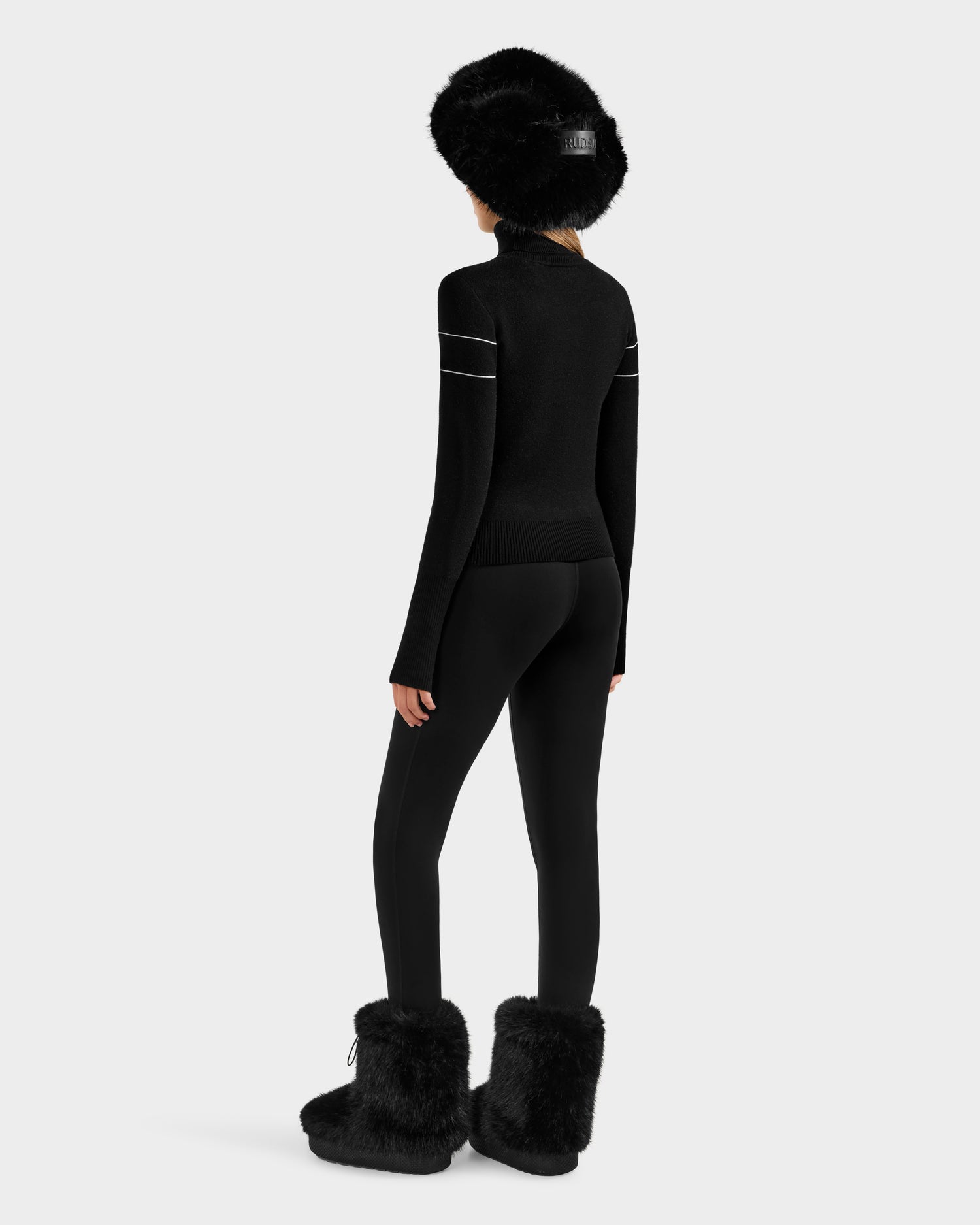 A person faces away, wearing the Rudsak ERIKA - 8523536 Black fuzzy boots, a black apres-ski turtleneck sweater with white stripes on the arms, black leggings, and a black fuzzy hat against a plain light background.