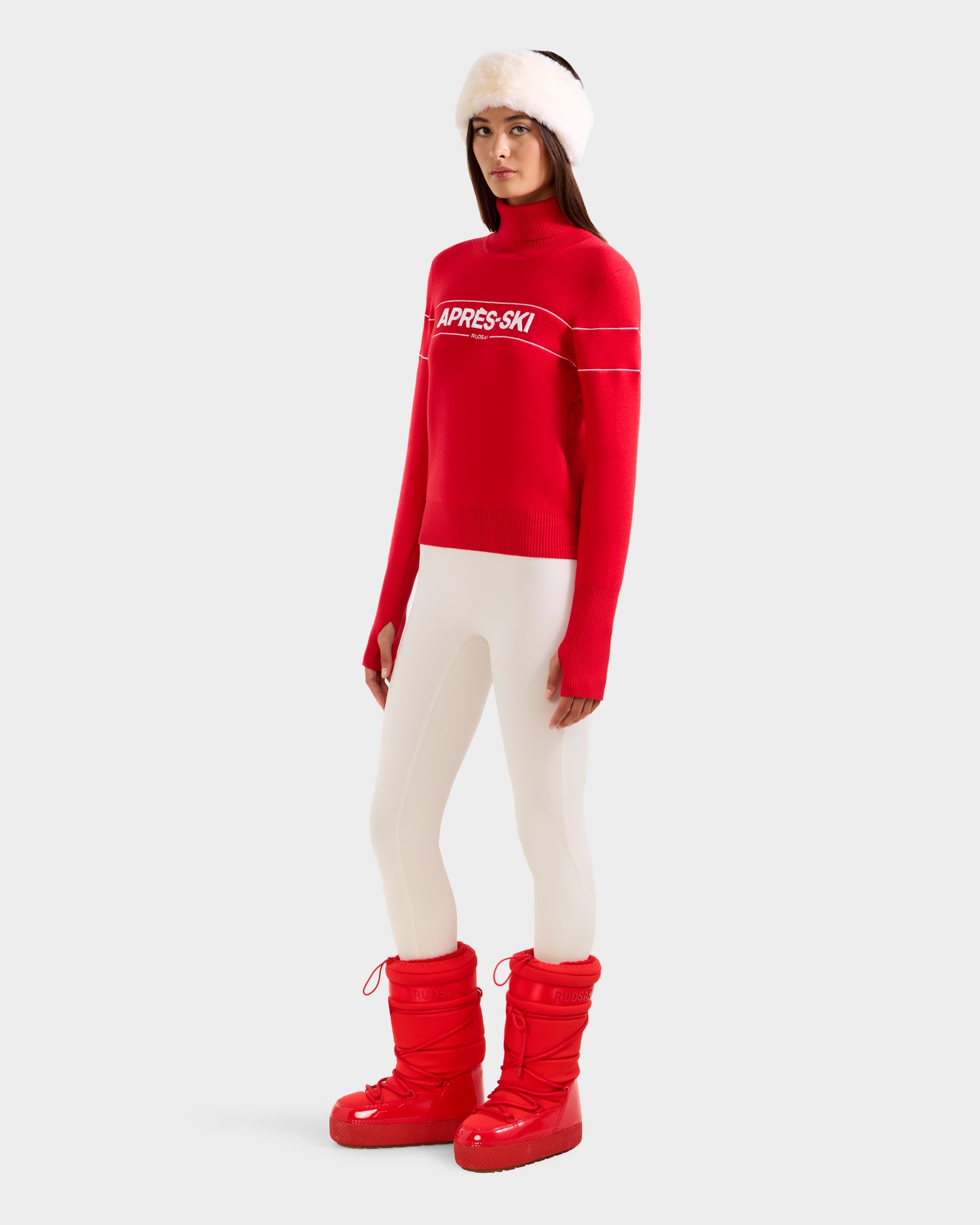 A woman in Rudsak's ERIKA - 8523536 Flame Red turtleneck, white fur hat, white leggings, and red snow boots stands before a light gray background—an effortless winter style statement.