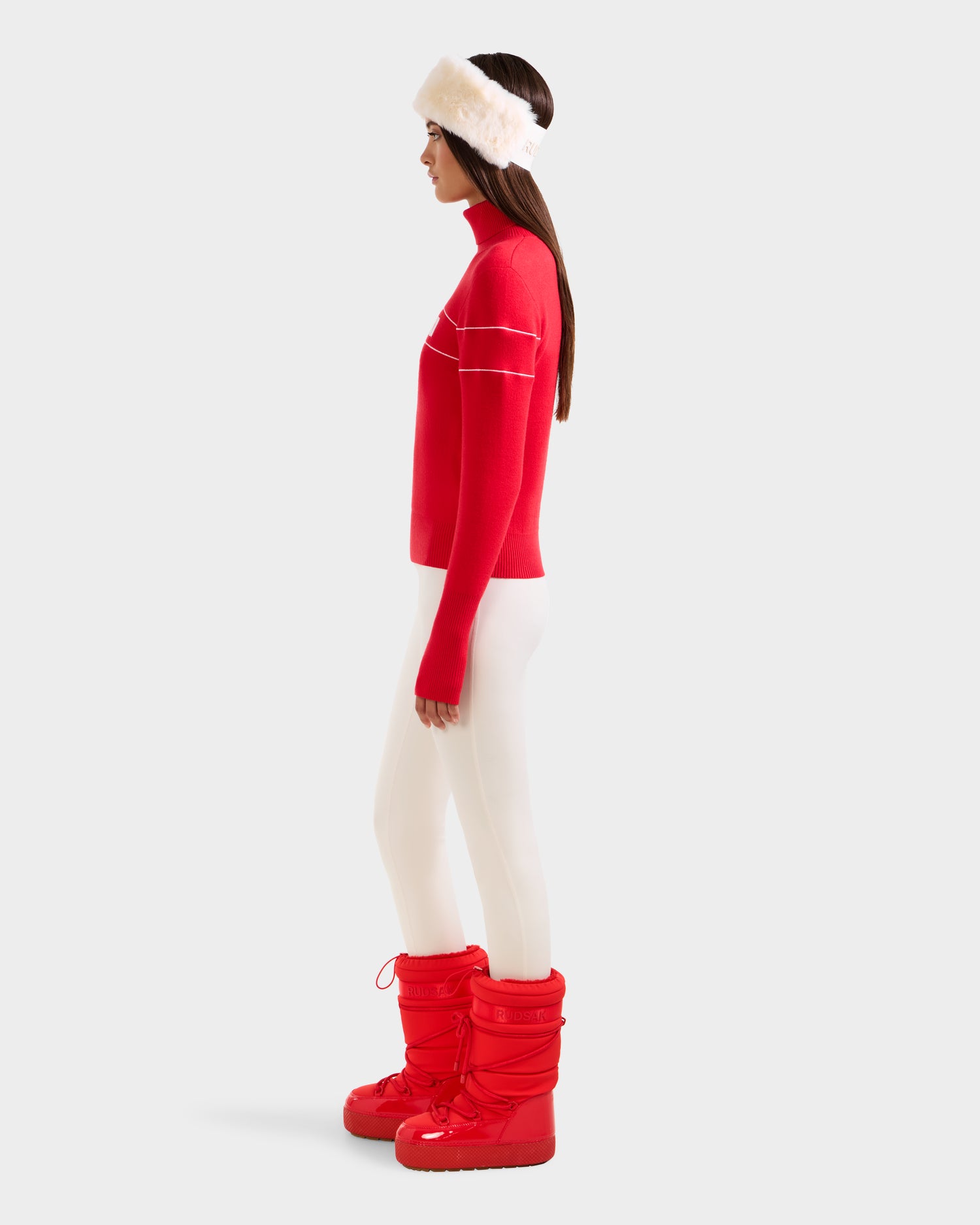 A woman in profile wears white leggings, a Rudsak après-ski turtleneck sweater, a white furry headband, and ERIKA - 8523536 Flame Red snow boots by Rudsak—an effortlessly stylish winter look set against a plain light gray background.