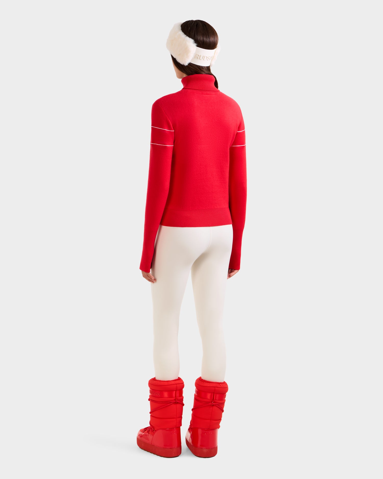 A woman faces away in Rudsak's ERIKA - 8523536 Flame Red turtleneck sweater, paired with white leggings, red puffer boots, and a white fuzzy headband against a plain light background, highlighting her chic winter style.