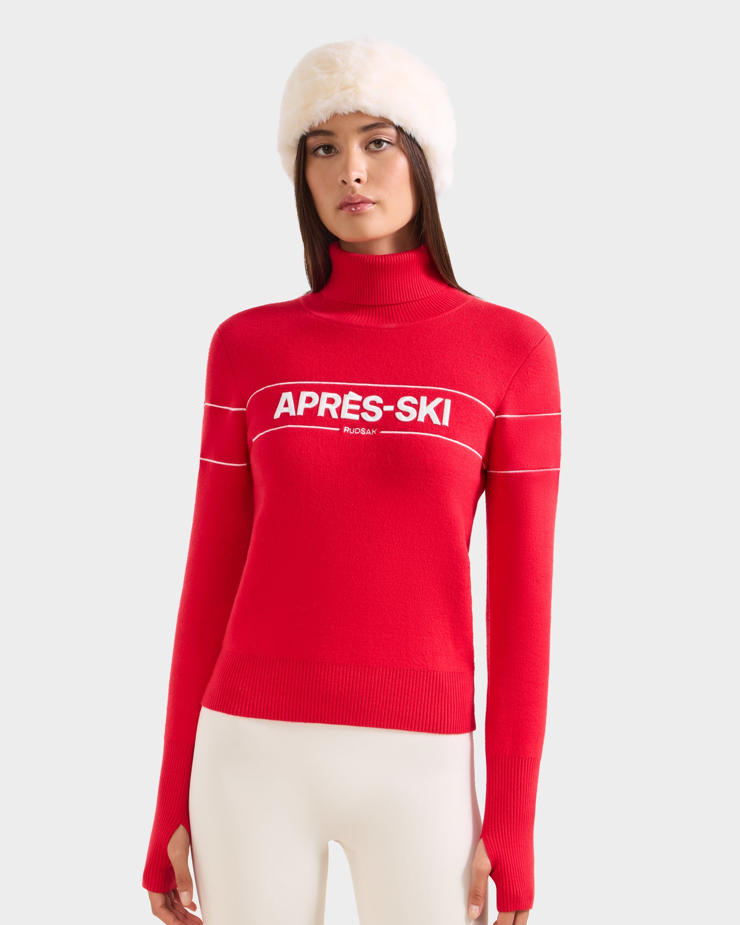 A woman in a white furry hat, white pants, and the Rudsak ERIKA - 8523536 Flame Red sweater with "APRÈS-SKI" on the chest stands against a light gray background, highlighting this chic winter staple.