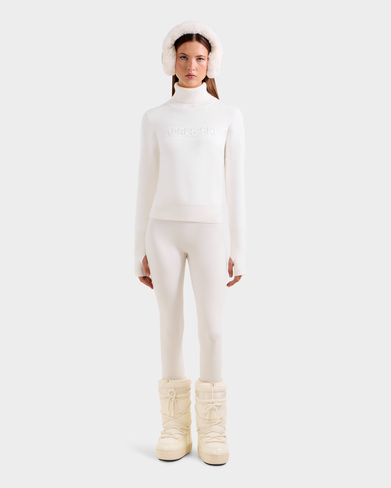 A woman faces forward against a plain light background, wearing the Rudsak ERIKA - 8523536 Iceland all-white apres-ski sweater with fitted pants, fluffy earmuffs, and padded snow boots.