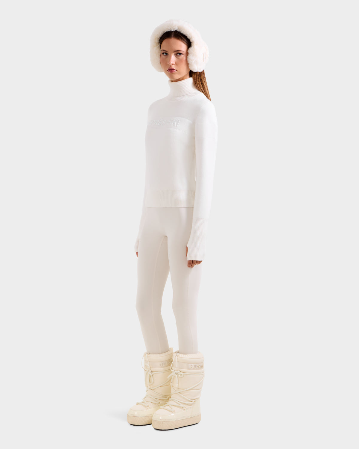 A woman stands against a light gray background wearing Rudsak’s ERIKA - 8523536 Iceland all-white outfit with a fur hat, après-ski turtleneck, fitted pants, and winter boots, looking forward with a neutral expression.