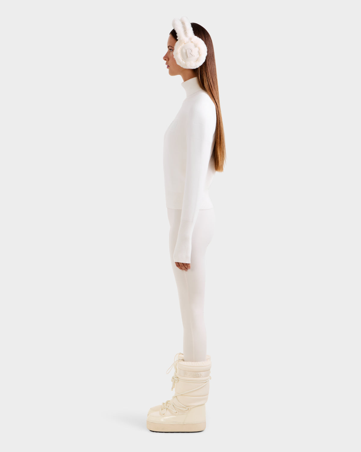 A woman in profile against a white background wears a white après-ski turtleneck, matching leggings, bunny ear fur earmuffs, and cream Rudsak ERIKA - 8523536 Iceland moon boots.