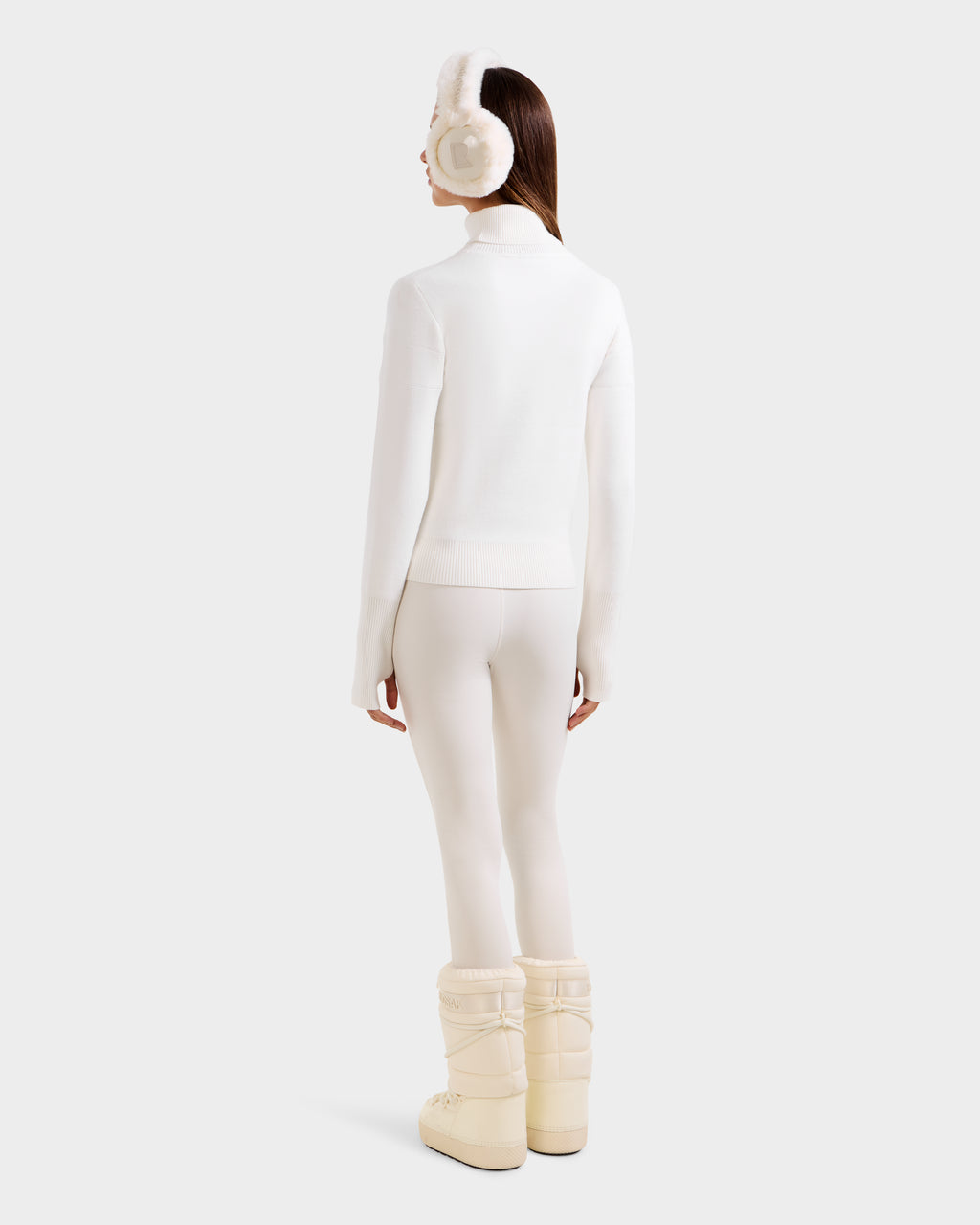 A person faces away, wearing the ERIKA - 8523536 Iceland après-ski sweater by Rudsak with matching leggings, earmuffs, and puffy boots against a plain light background.