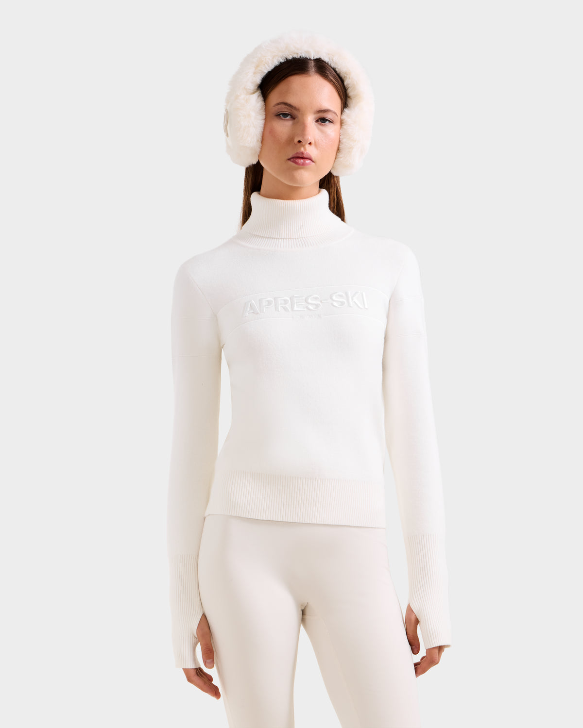 A woman stands against a plain light background wearing the Rudsak ERIKA - 8523536 Iceland turtleneck sweater with white pants and fluffy earmuffs—a chic, effortless addition to your winter wardrobe.