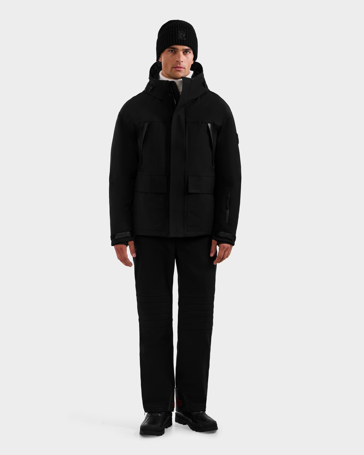 A person faces forward in a Rudsak ETHAN - 6125589 Black men’s ski jacket, black pants, shoes, and knit beanie, set against a plain light gray background.