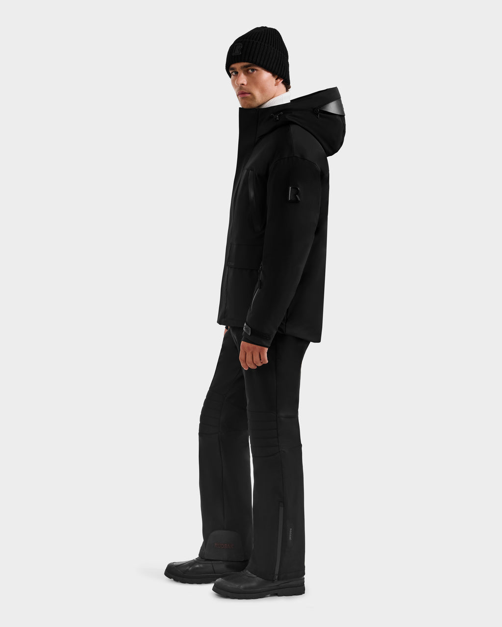 Wearing the Rudsak ETHAN - 6125589 Black men's ski jacket with a hood, black pants, boots, and a knit beanie, a person stands against a light gray background, looking slightly to the side.