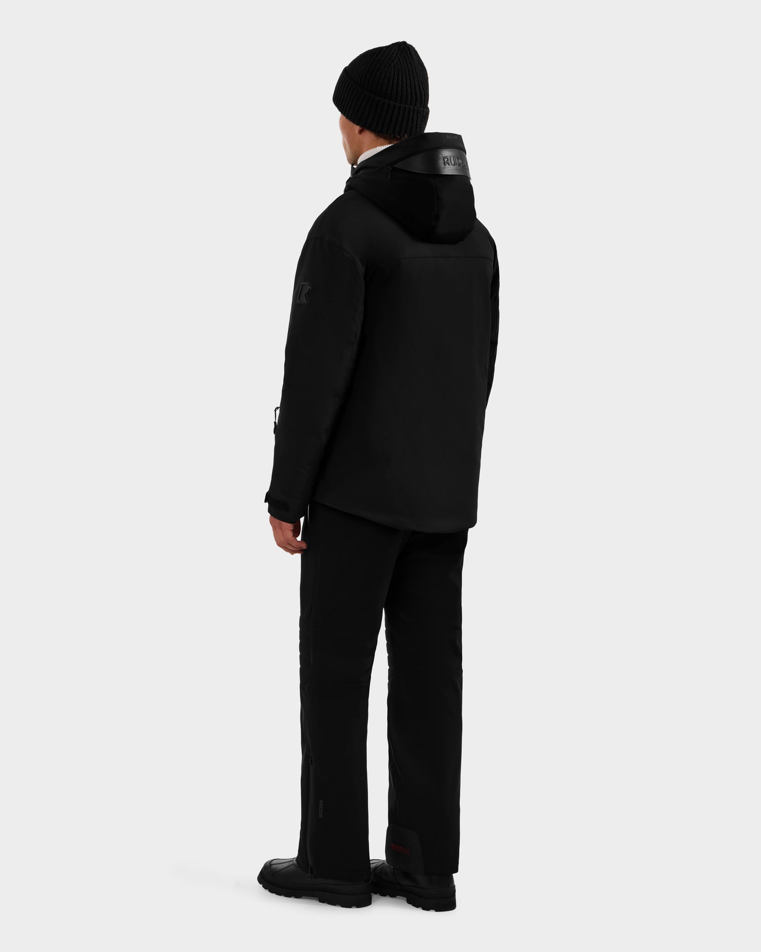 A person wearing the ETHAN - 6125589 Black men’s ski jacket by Rudsak, along with a black beanie, pants, and boots, stands facing away from the camera against a plain light gray background.