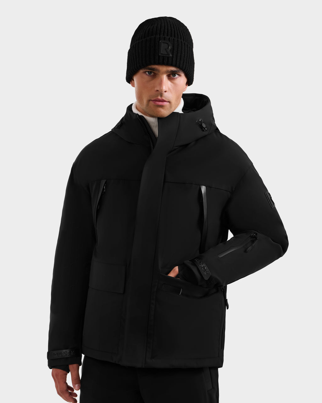 A man stands against a plain light gray background, looking straight ahead with one hand in his pocket, wearing the Rudsak ETHAN - 6125589 Black men’s ski jacket.