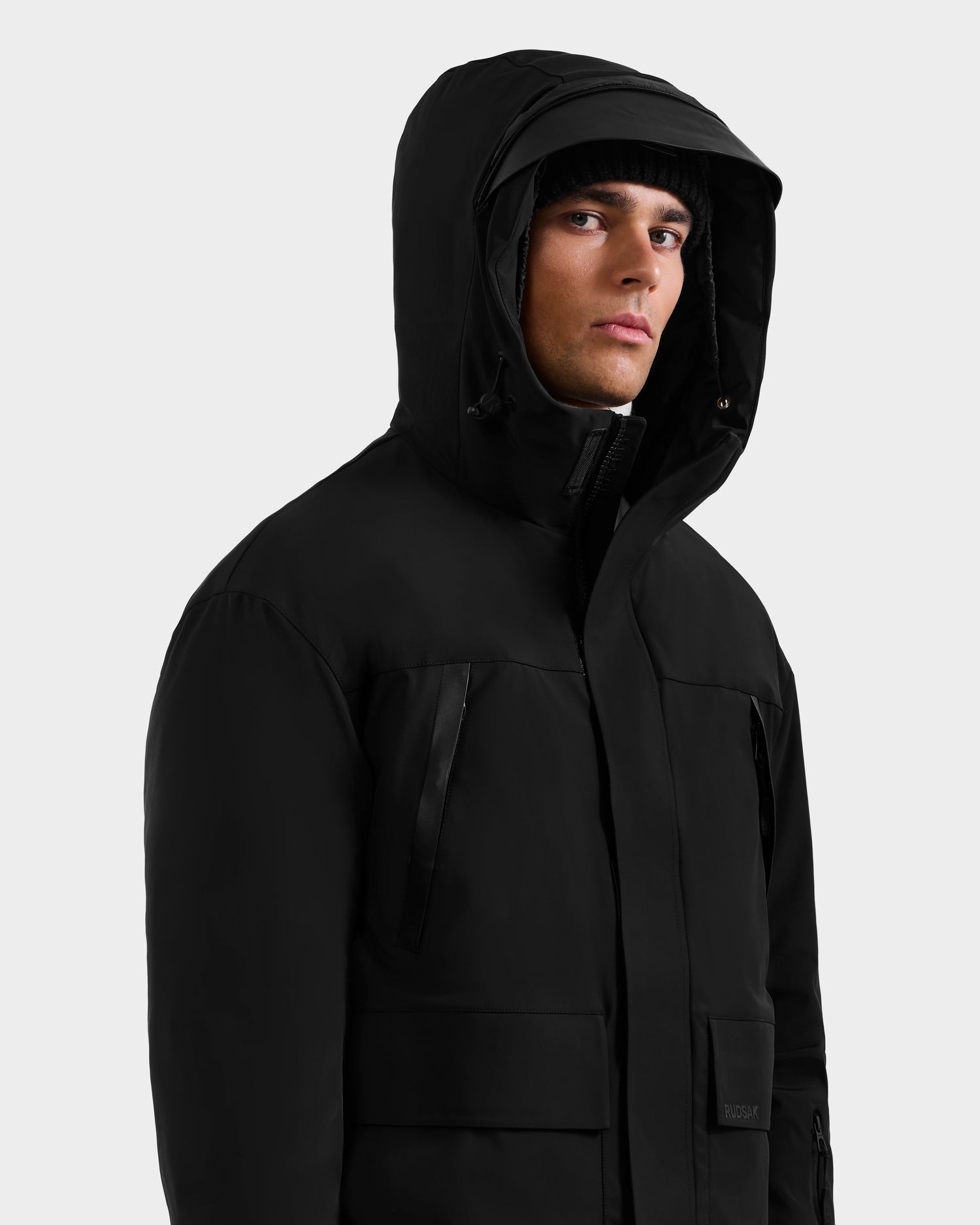A person wearing Rudsak's ETHAN - 6125589 Black men’s ski jacket with RDS-certified down and a black beanie, looking slightly to the side against a plain light background.