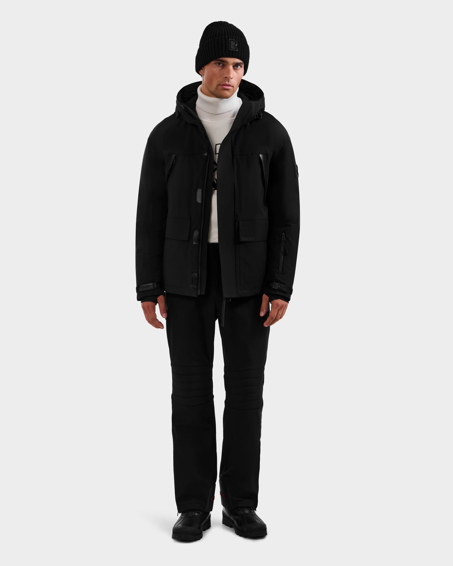 A man faces forward in a plain light gray setting, wearing the Rudsak ETHAN - 6125589 Black men’s ski jacket, paired with black beanie, pants, and shoes over a white turtleneck sweater.