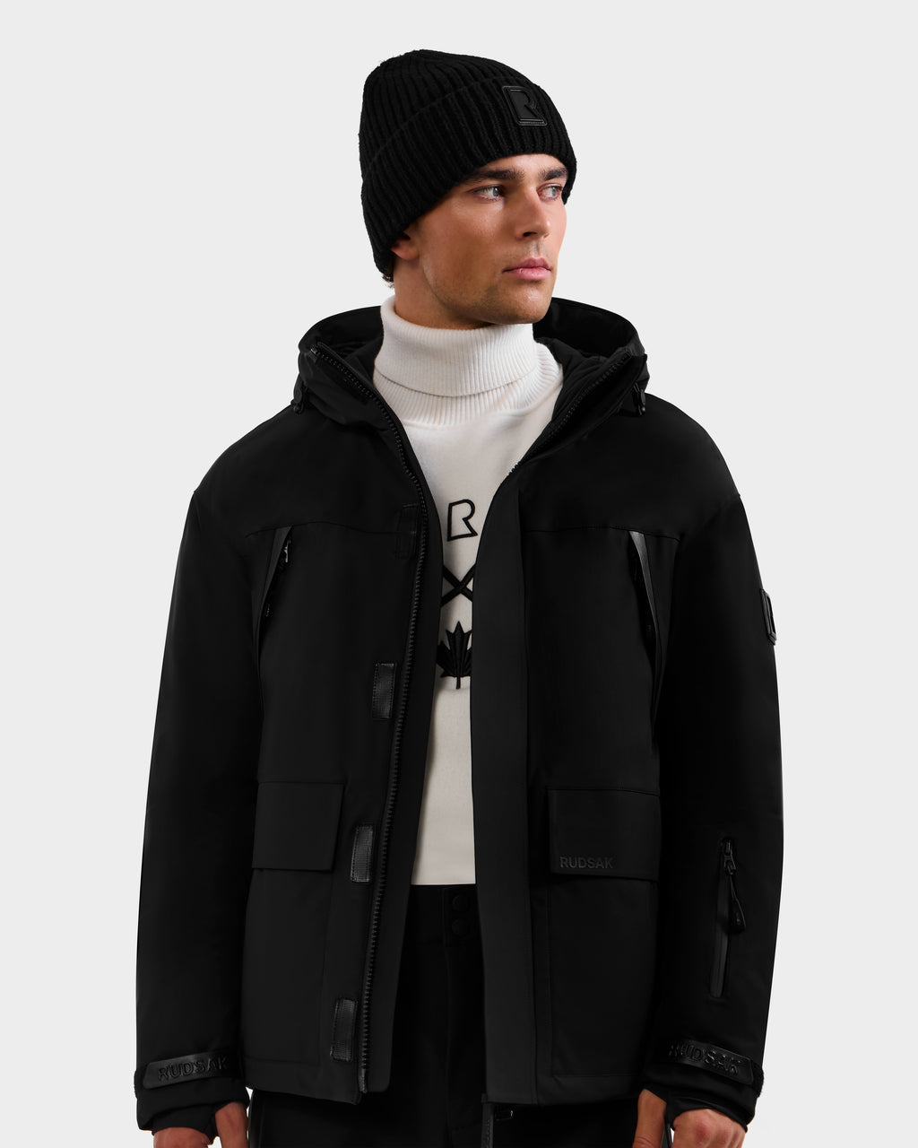 A man wearing the Rudsak ETHAN - 6125589 Black men’s ski jacket stands against a plain light gray background, styled with a black beanie and a white turtleneck sweater featuring a black geometric design.