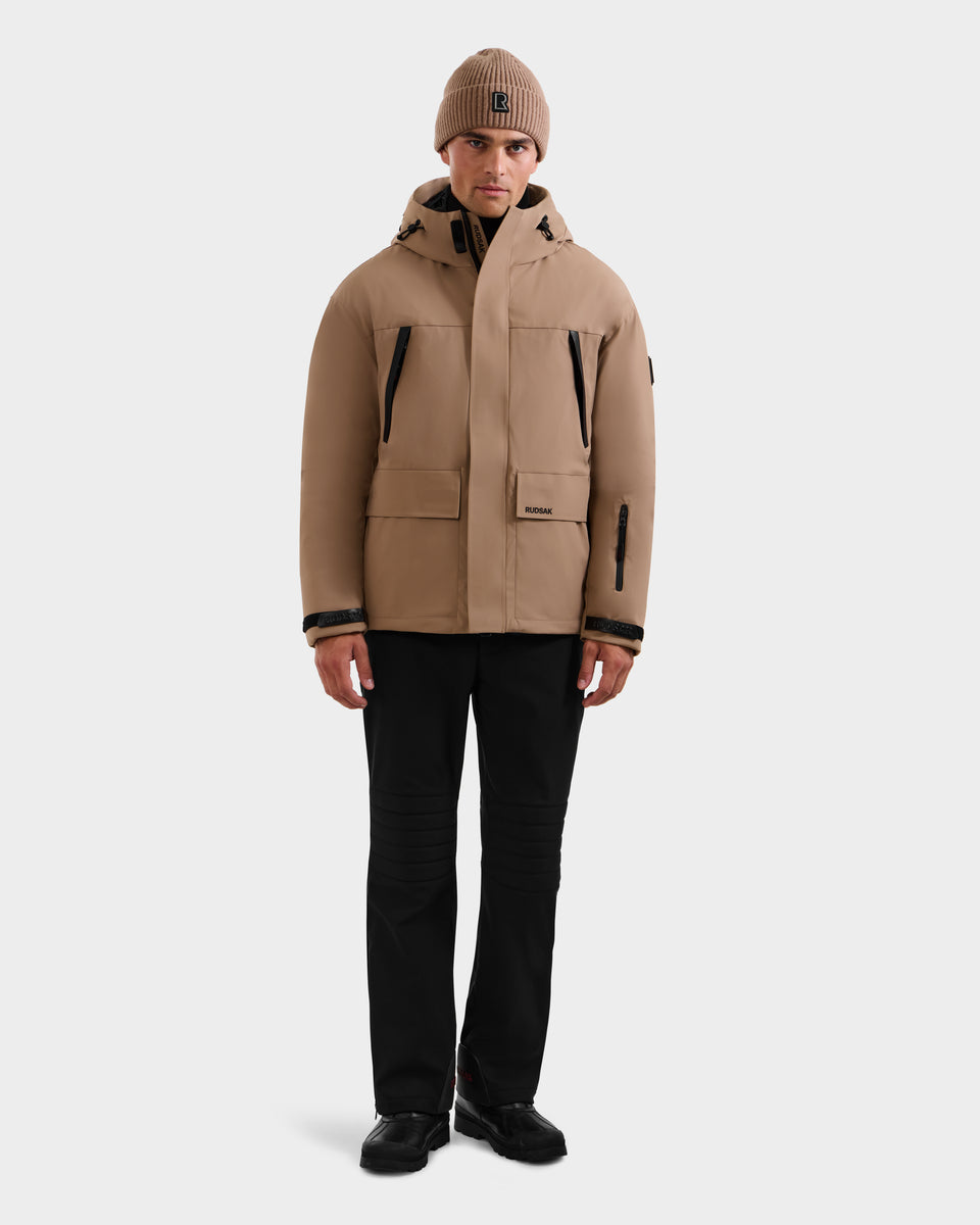 A man faces forward wearing the Rudsak ETHAN - 6125589 Sand men's ski jacket with large pockets, a matching tan beanie, black pants, and black shoes against a plain light background.