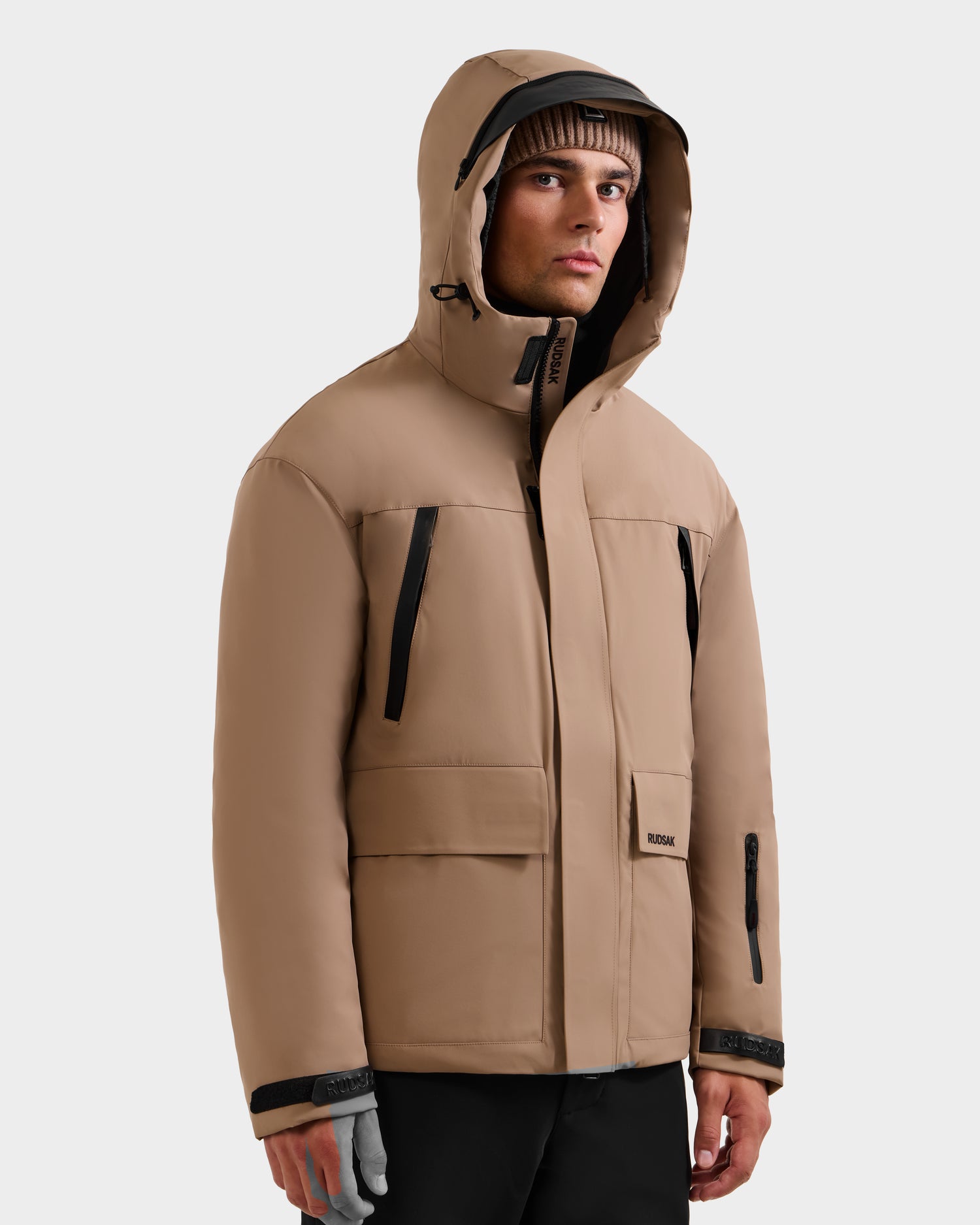 A man in the Rudsak ETHAN - 6125589 Sand men's ski jacket with black zippers and a brown beanie stands against a plain light background, looking slightly to the side.