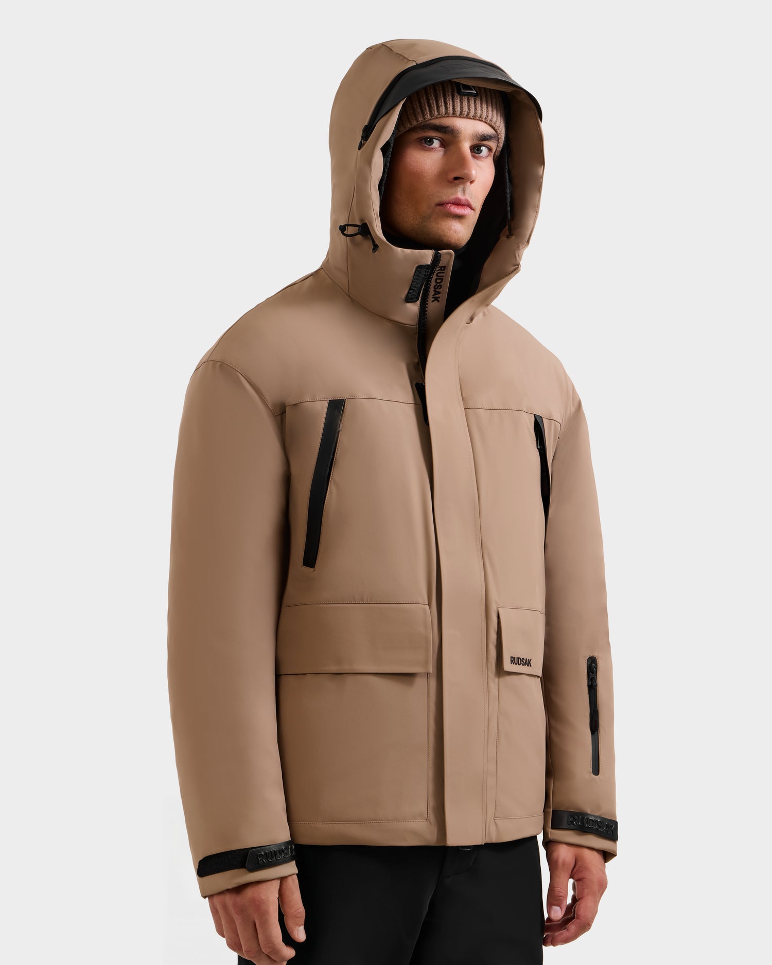 A man stands against a plain light background, looking slightly to the side, wearing the Rudsak ETHAN - 6125589 Sand men’s ski jacket with black zippers and a brown beanie.