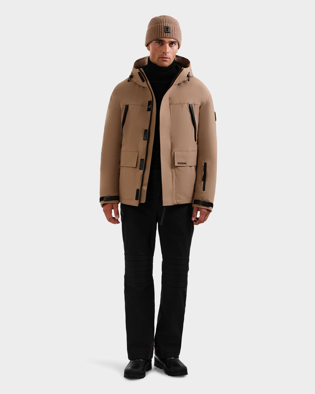 A person wearing the Rudsak ETHAN - 6125589 Sand men's ski jacket, a matching tan beanie, black pants, and black shoes stands facing forward against a plain white background.