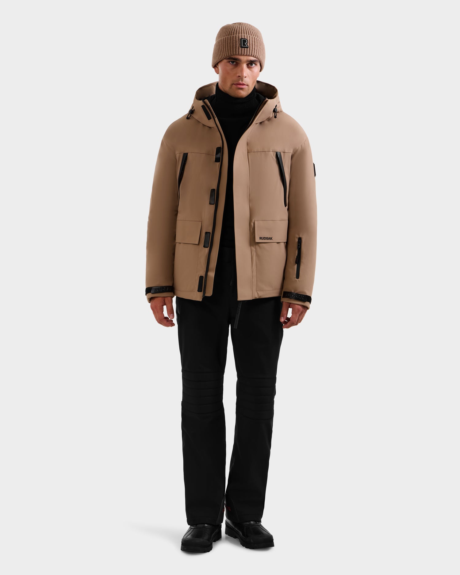 A person wearing the Rudsak ETHAN - 6125589 Sand men's ski jacket, a matching tan beanie, black pants, and black shoes stands facing forward against a plain white background.