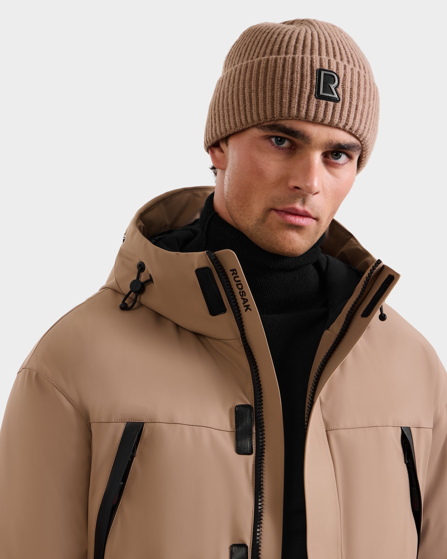 A man wears Rudsak's ETHAN - 6125589 Sand ski jacket with black zippers and RDS-certified down, paired with a matching beige Rudsak knit beanie over a black turtleneck, shown against a plain background.