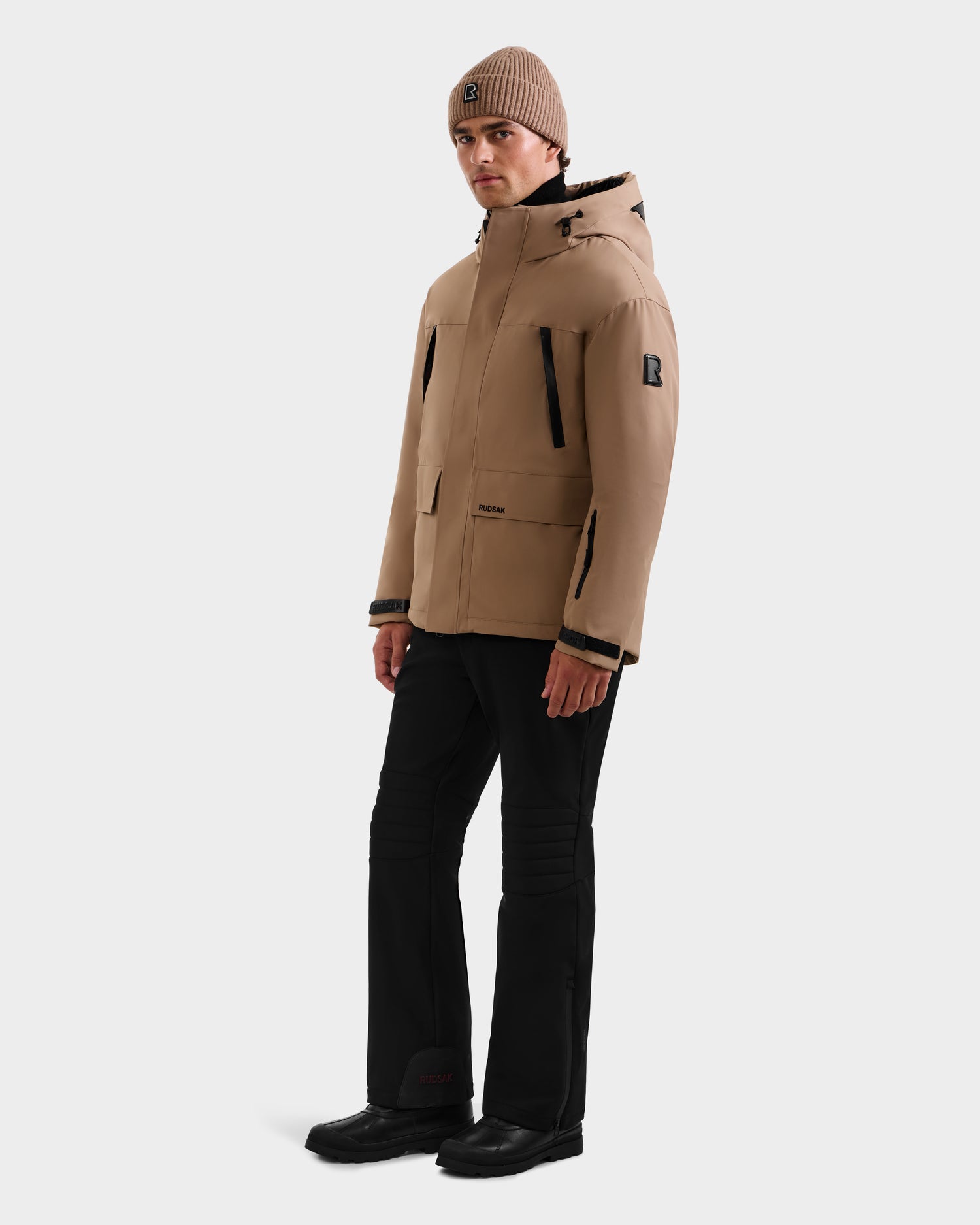 A person wearing the Rudsak ETHAN - 6125589 Sand men's ski jacket with hood, a matching tan beanie, black pants, and black shoes stands against a plain light background.