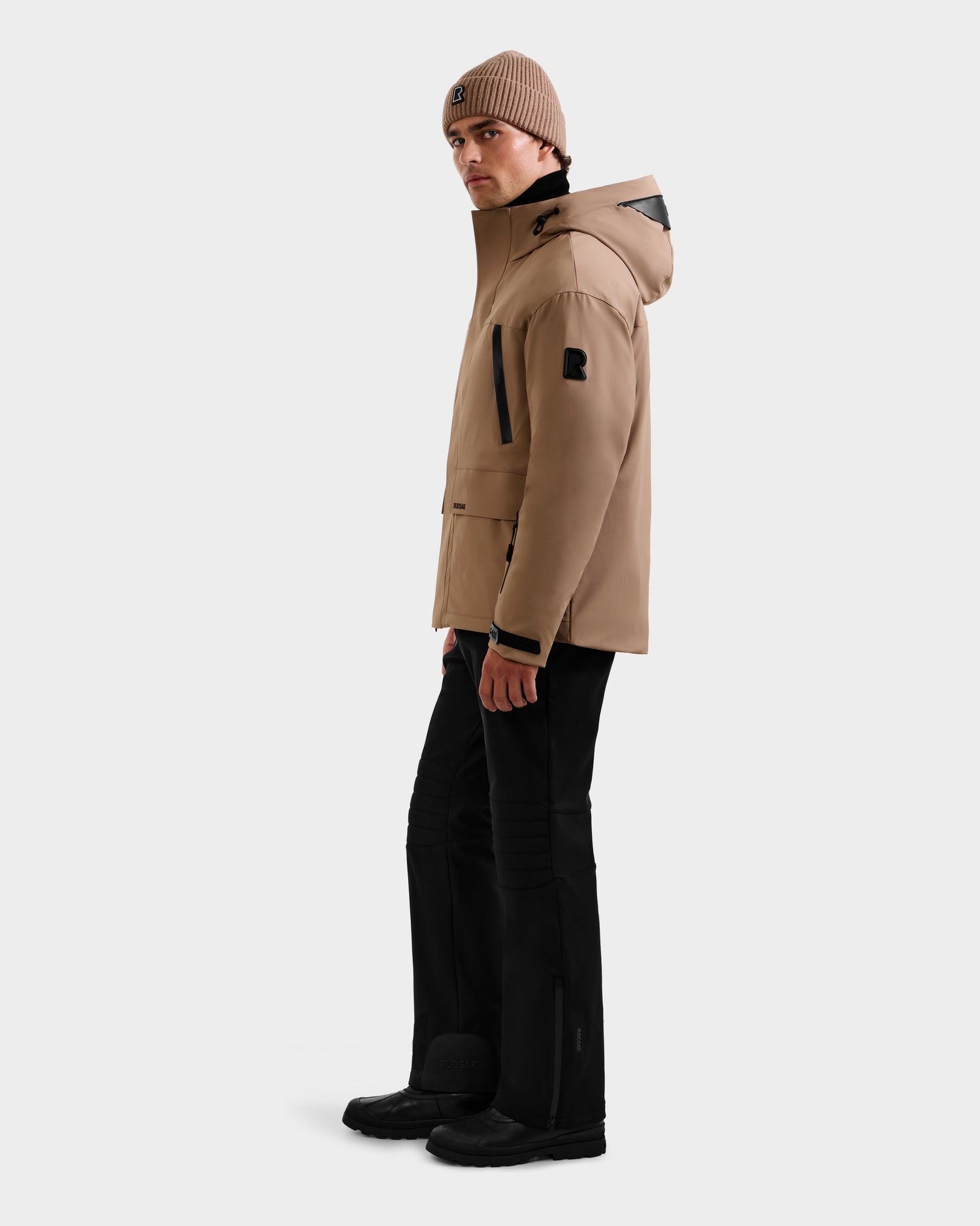 A person wears the Rudsak ETHAN - 6125589 Sand RDS-certified beige down jacket, black pants and shoes, with a beige knit beanie, standing sideways and looking slightly toward the camera against a plain light gray background.