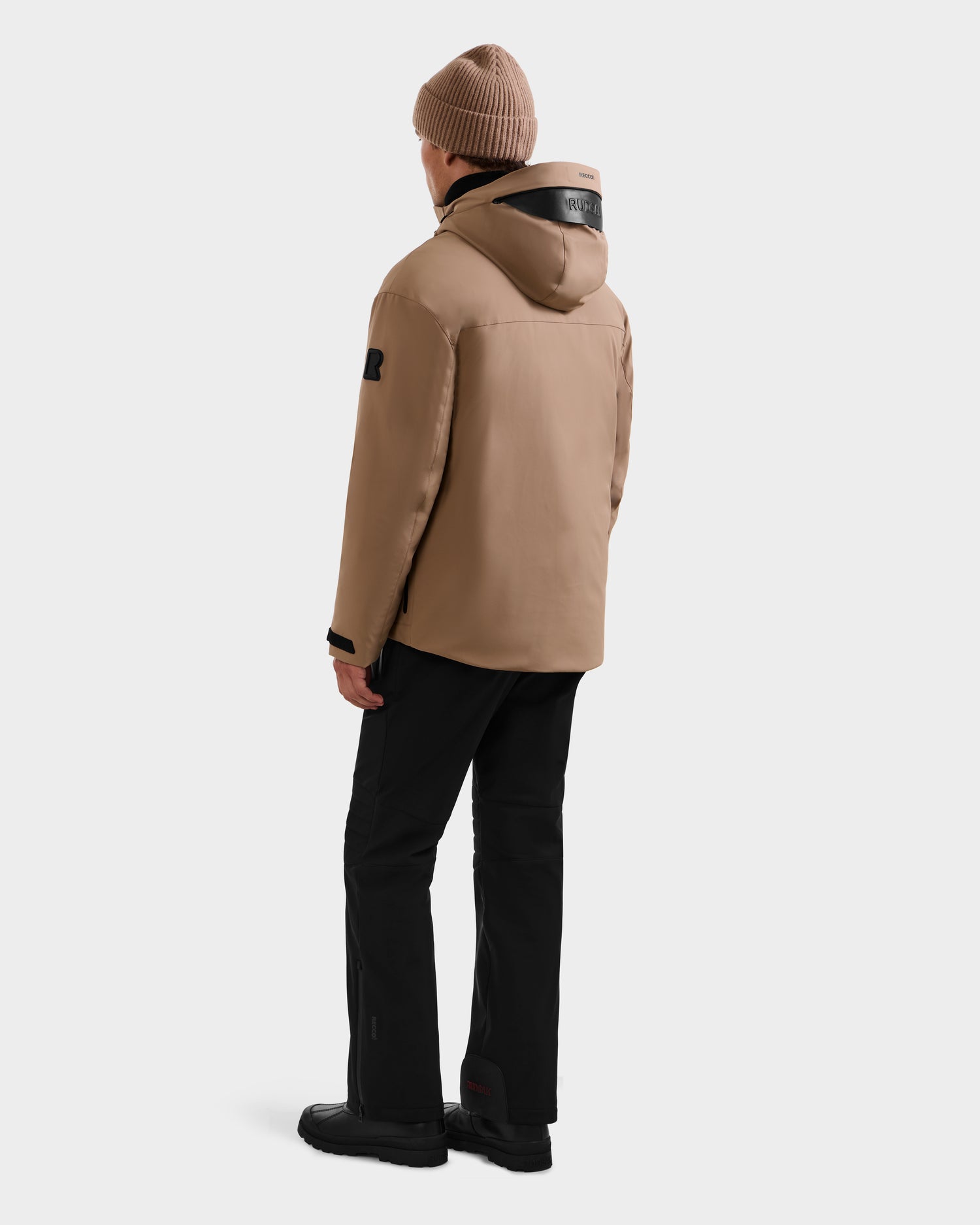 A person stands facing away against a plain white background, wearing Rudsak's ETHAN - 6125589 Sand jacket with black pants, black shoes, and a brown knit beanie.
