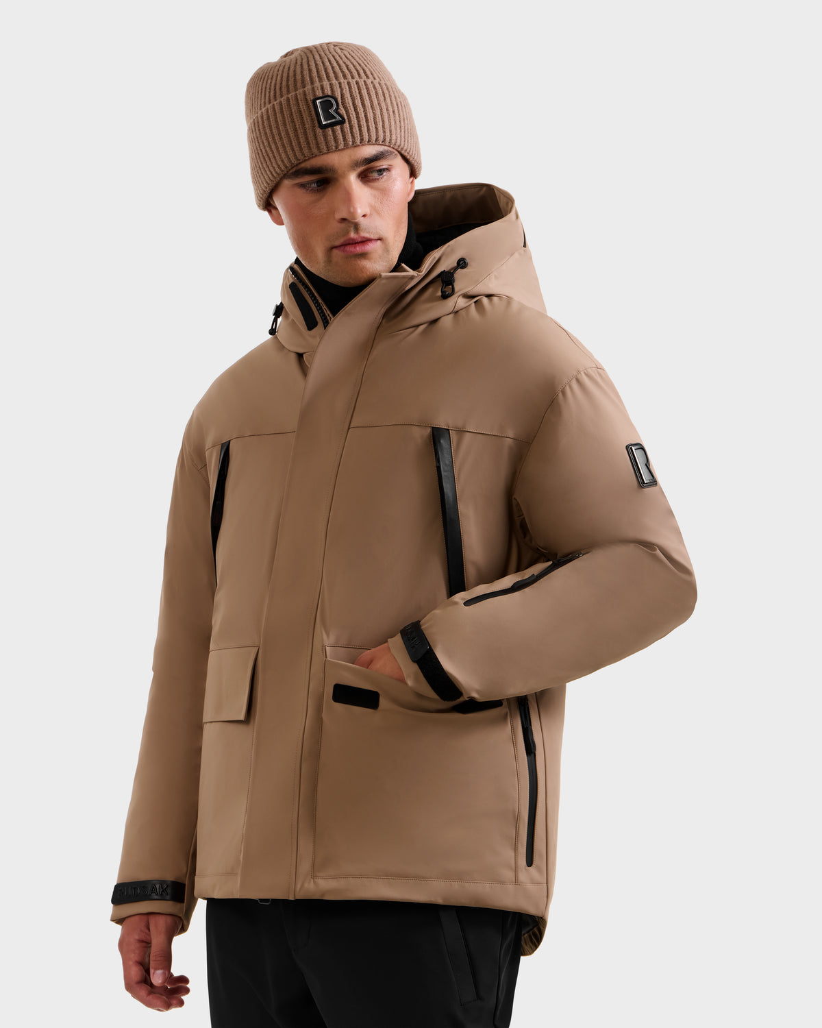 A man wears the Rudsak ETHAN - 6125589 Sand men’s ski jacket with black zippers and a matching tan beanie, both featuring a black 'R' logo, against a plain light background. The jacket is filled with RDS-certified down for warmth and comfort.