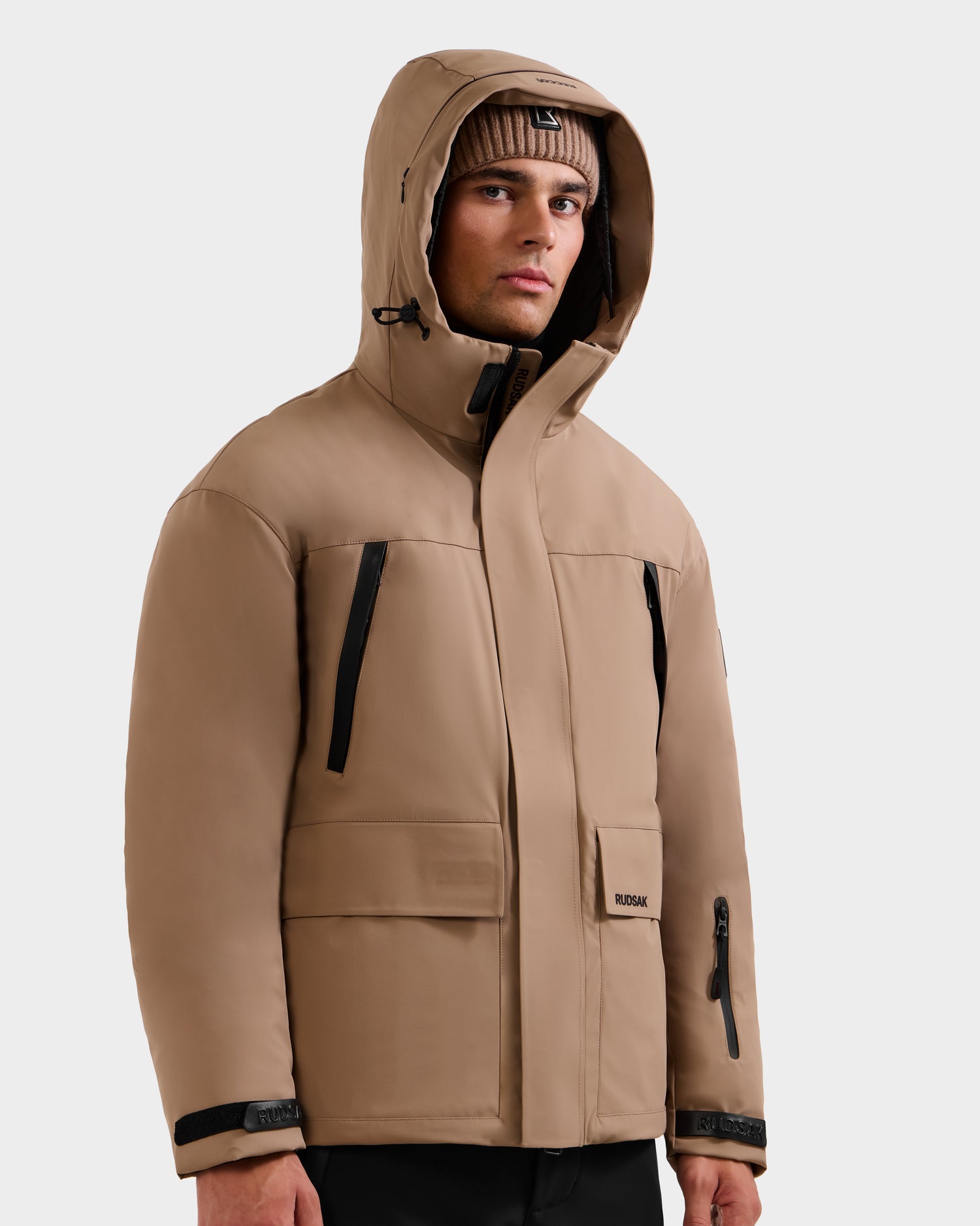 A man wears the Rudsak ETHAN - 6125589 Sand men’s ski jacket with black zippers and flap pockets, paired with a matching knit beanie, standing slightly to the side against a plain light background.