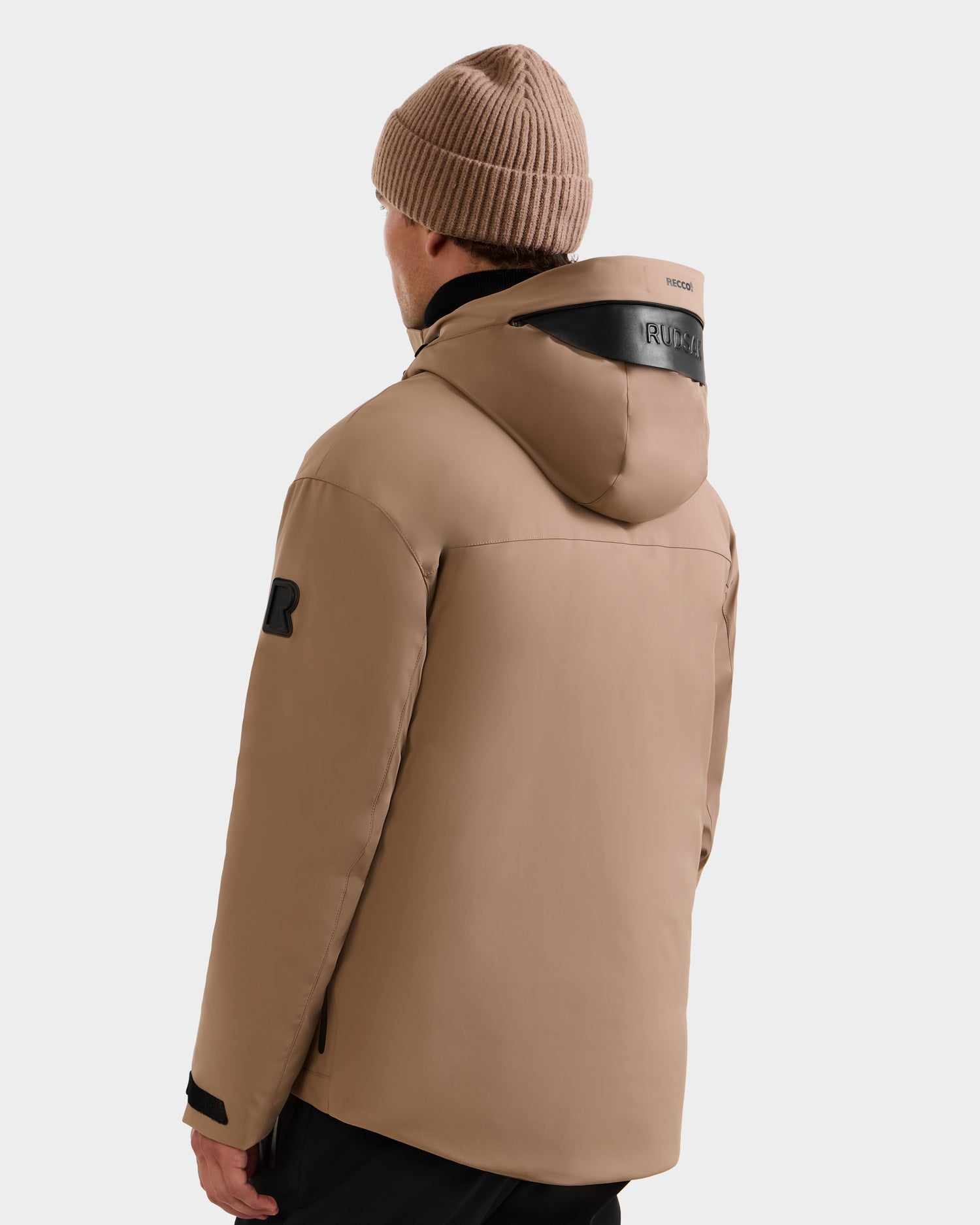 A person wearing the ETHAN - 6125589 Sand men's ski jacket by Rudsak and a brown knit beanie faces away, highlighting the back and hood details of the jacket against a plain white background.
