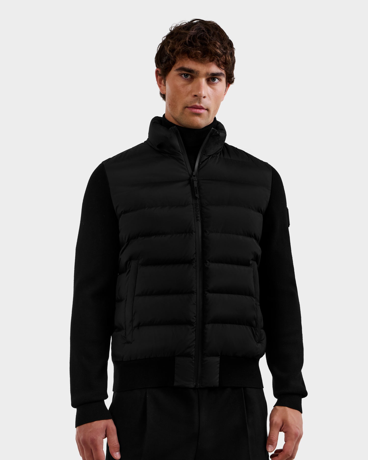 A man with short brown hair wears the Rudsak EUGENE - 6124721 Black performance jacket over a black long-sleeve shirt and black pants, standing against a plain light gray background.