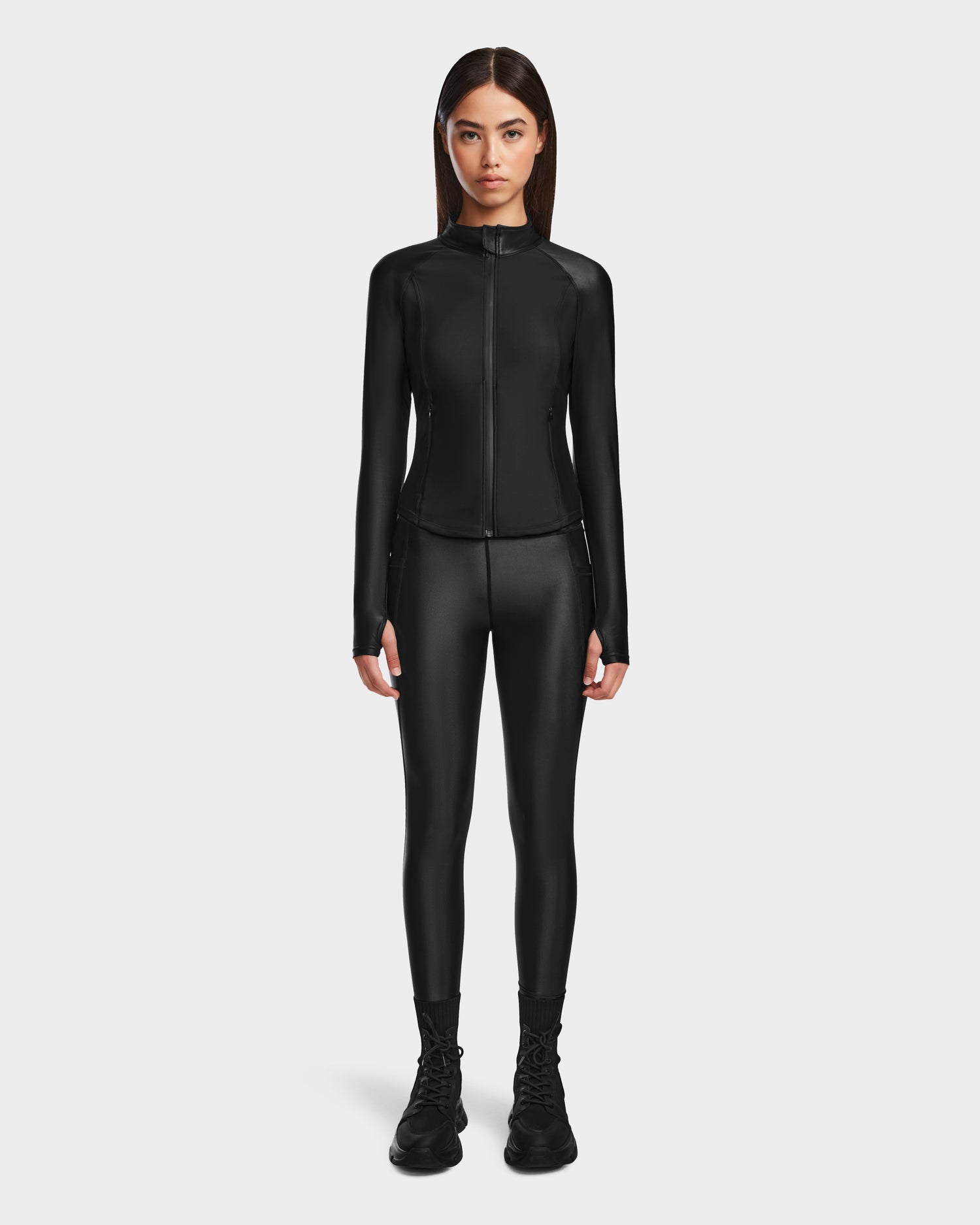 A woman faces forward against a white background, wearing the Rudsak EVA - 8524514 Black zip-up top with matching black leggings and boots, all in shiny stretch fabric.