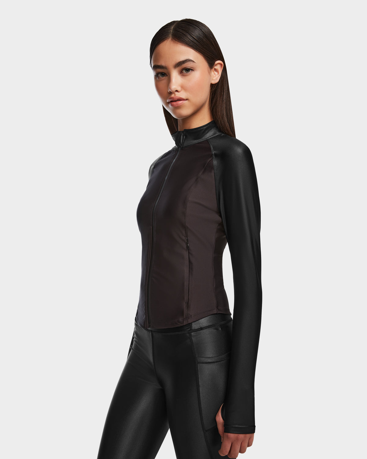 A woman with long dark hair wears the Rudsak EVA - 8524514 Black fitted womens zip-up top and black pants, standing confidently against a white background.