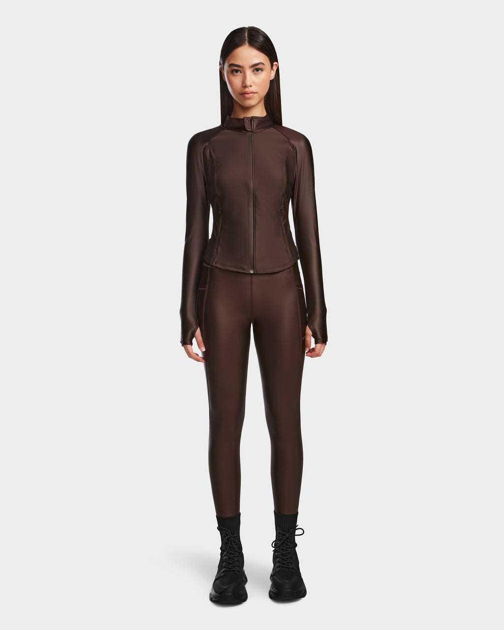 A woman faces forward wearing the Rudsak EVA - 8524514 Espresso fitted zip-up top and matching leggings with a waterproof zipper and black boots, set against a plain white background.
