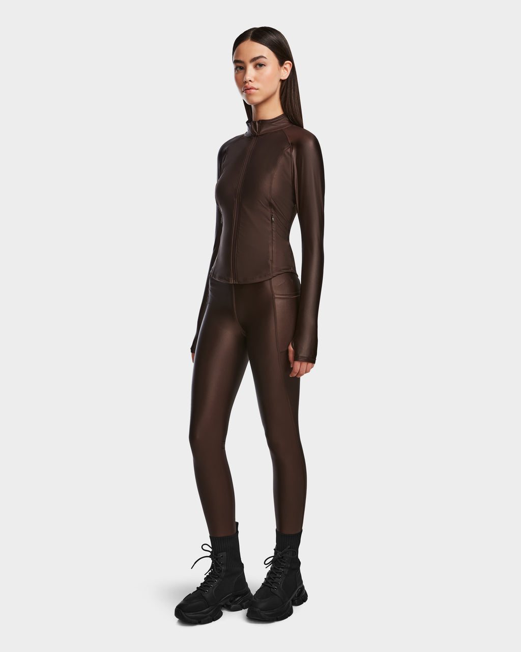 A woman with long straight hair and a neutral expression stands against a light background, wearing the Rudsak EVA - 8524514 Espresso fitted zip-up top and matching leggings in shiny stretch fabric, paired with black boots.