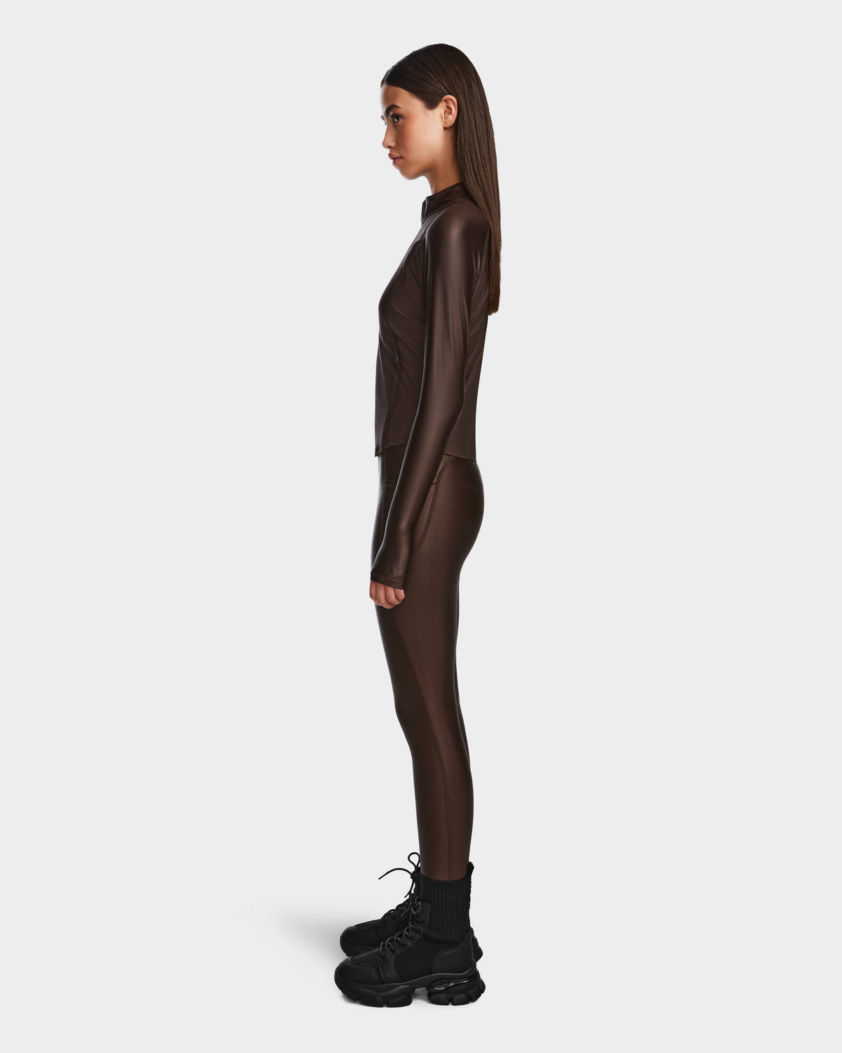 A woman stands in profile against a white background, wearing the Rudsak EVA - 8524514 Espresso fitted zip-up top and matching pants in shiny stretch fabric with a waterproof zipper, paired with black chunky boots.